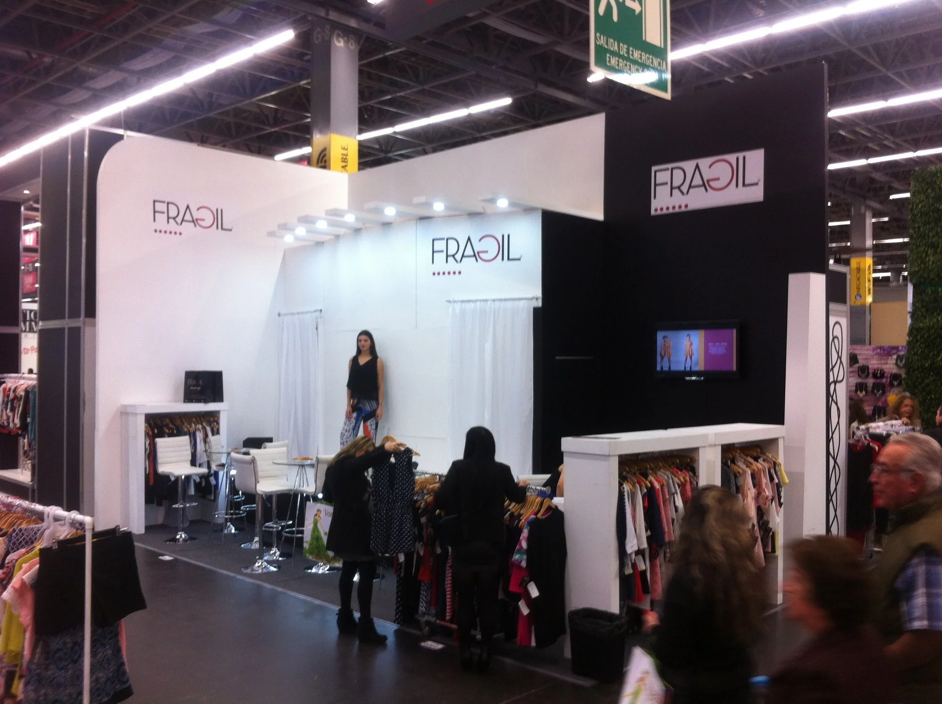 Stand de ropa de marca Fragil en una feria, con una mujer en el escenario y varias personas alrededor mirando la ropa.