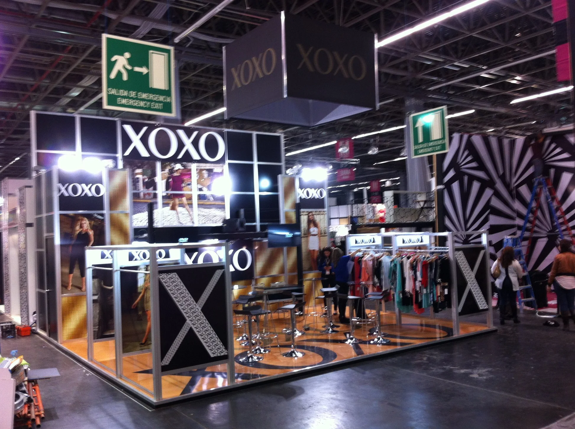 Stand de la marca XOXO en una feria, decorado en negro y dorado, con exhibición de ropa y pantallas mostrando modelos