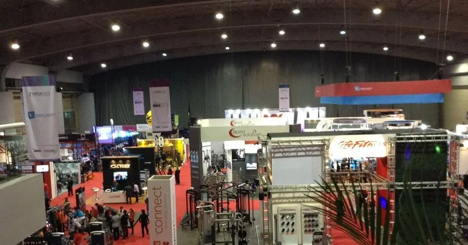 Vista de un evento o feria en un espacio cerrado con stands y exhibiciones, con personas recorriendo y interactuando. Iluminación artificial y decoración variada.