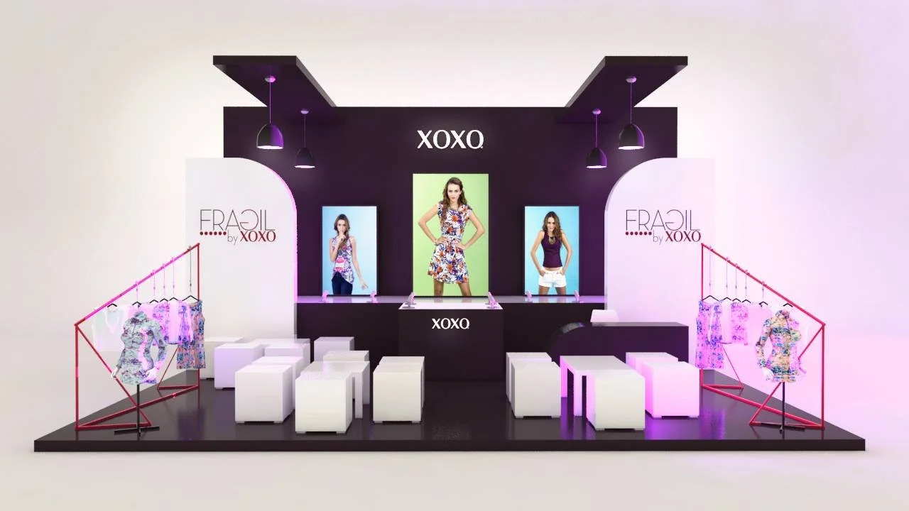 Stand de exhibición de moda con fondos de pantalla con la marca 'XOXO', exhibiendo modelos con ropa femenina y accesorios, con escritorios blancos y bancos para visitantes.