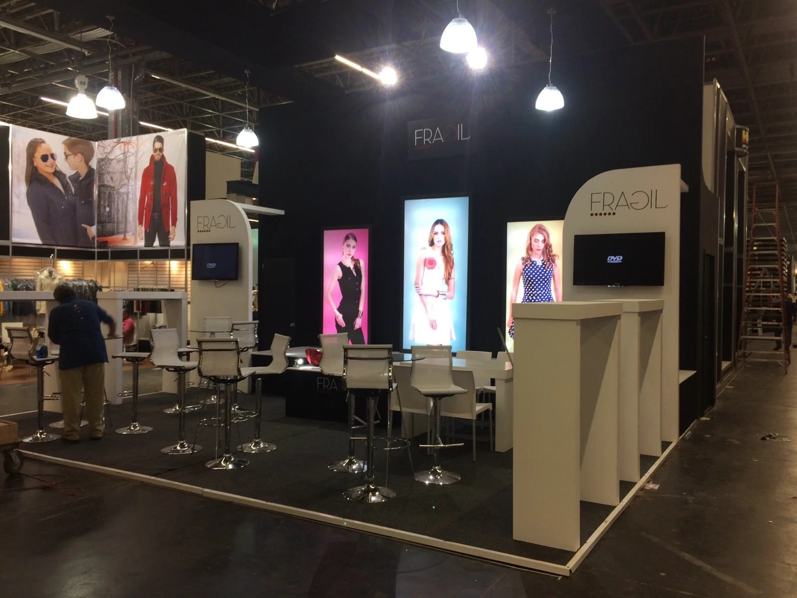Stand de exhibición con pantallas y sillas en un evento, con carteles de modelos y el texto 'FRAIL' en la parte superior.