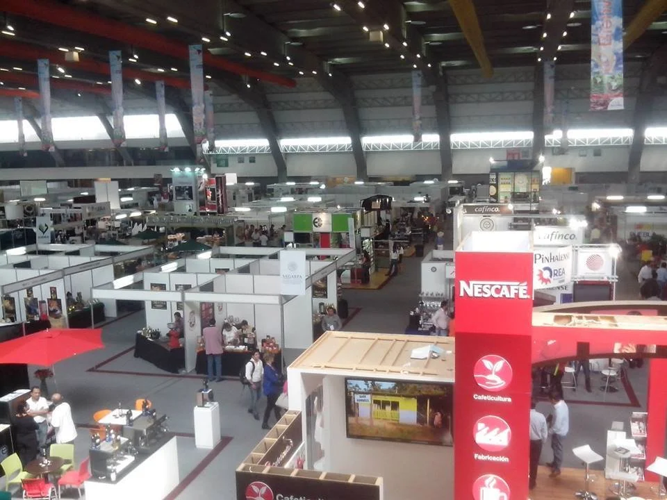 Vista de un centro de exposiciones con varias casetas y puestos comerciales, algunas con personas comprando y caminando.