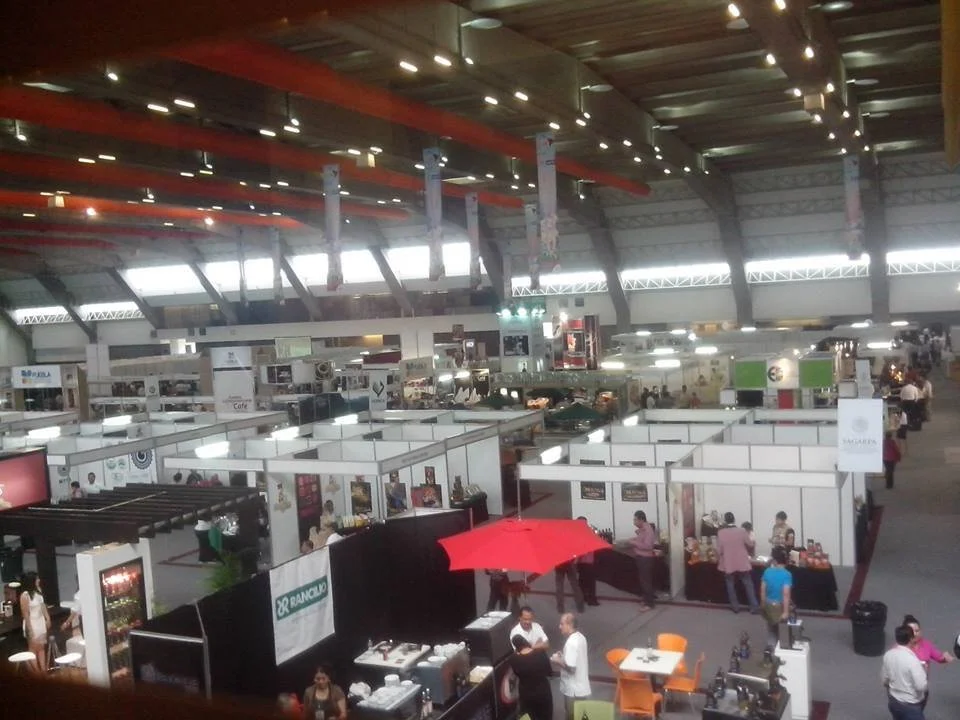Vista de un salón de exhibiciones con varias casetas y puestos, gente caminando y trabajando, y un techo con luces y vigas.