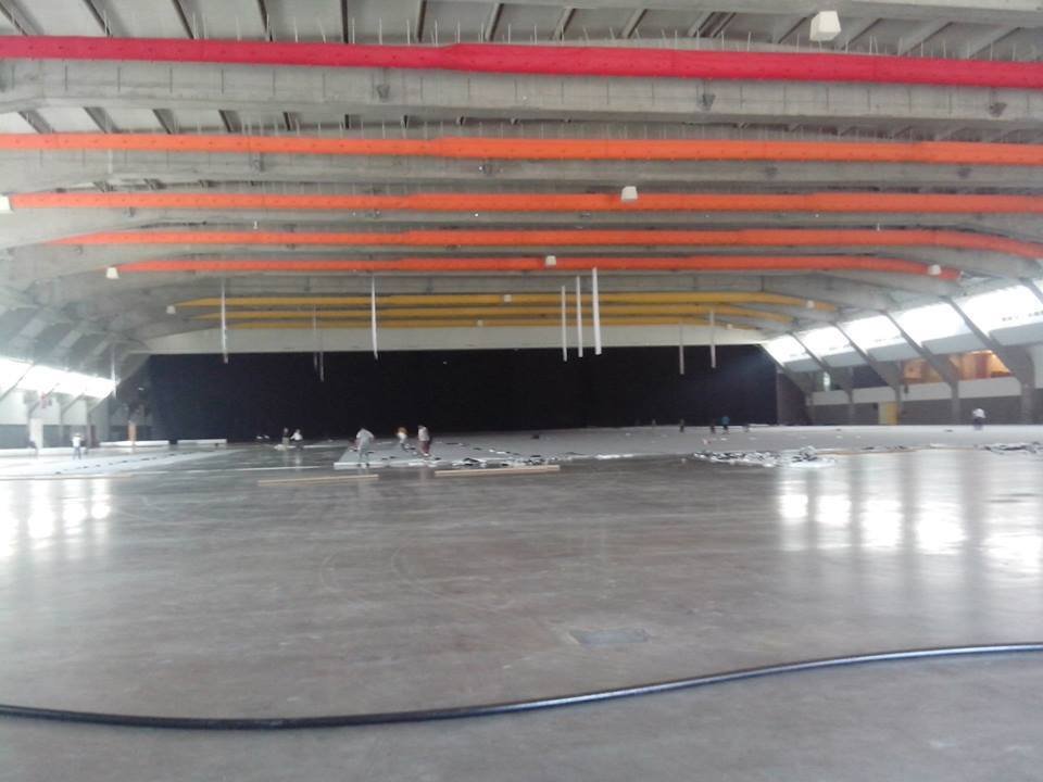 Interior de un estadio grande, en construcción o reparación, con estructura de techo visible y varias personas trabajando en el suelo.