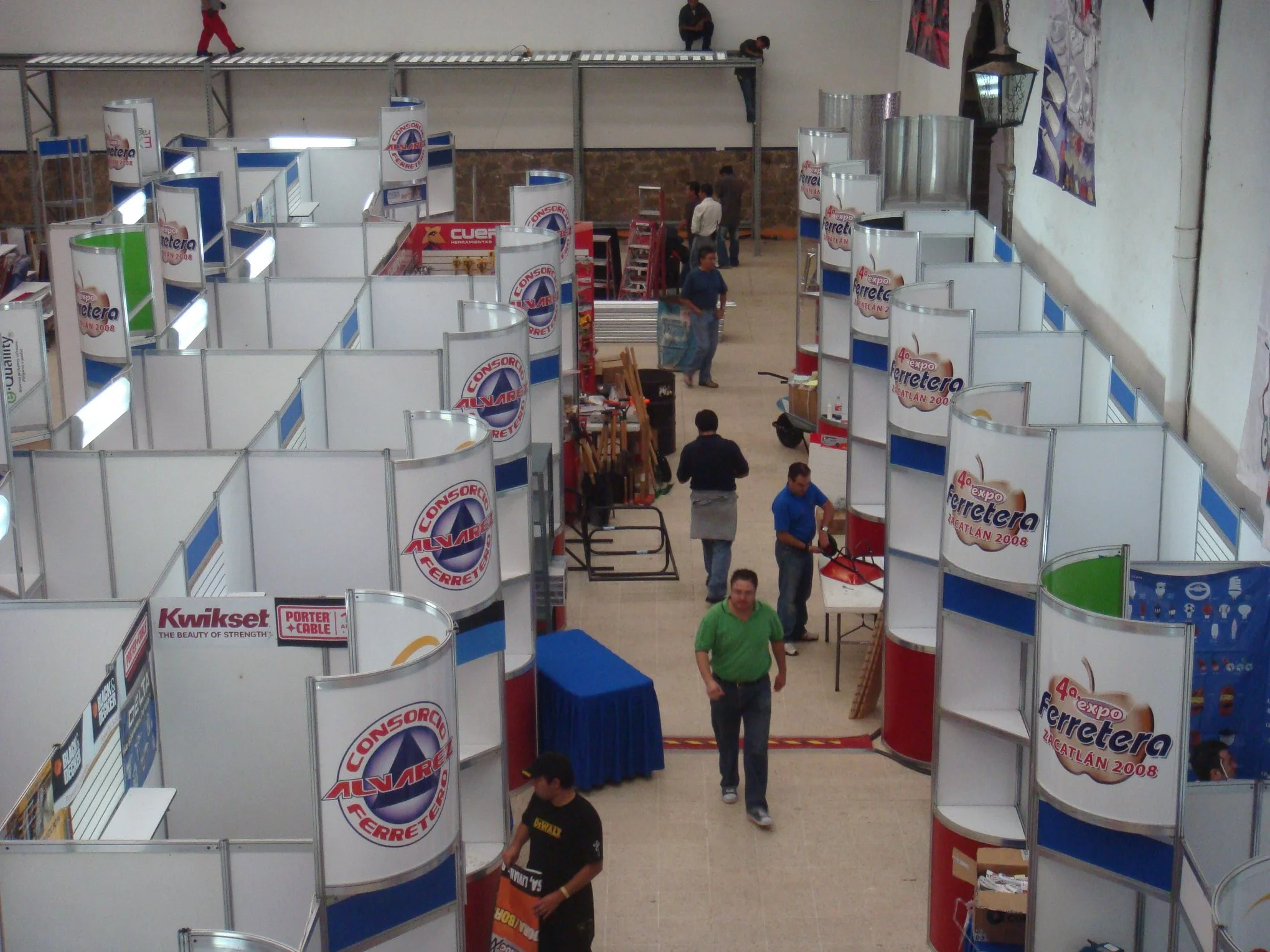Vista aérea de un pabellón de feria con varios puestos y cabinas en montaje, y algunas personas caminando y preparando los stands.