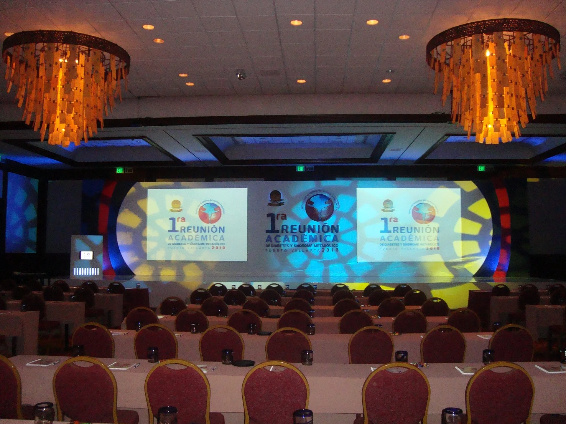 Salón de conferencias preparado para una reunión académica sobre diabetes y síndrome metabólico, con varias sillas y mesas, y pantallas de proyección en el escenario mostrando el evento.