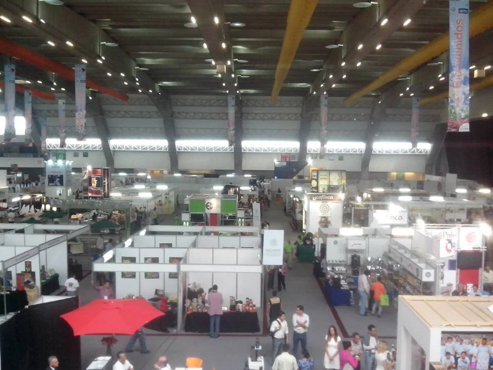 Vista general de feria comercial con puestos y visitantes en un recinto amplio y techado