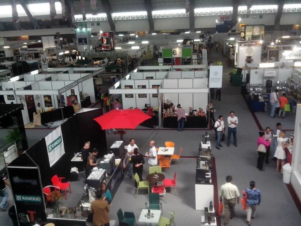 Imagen de un evento o feria en un recinto grande con diversas estaciones y puestos, algunas con exhibiciones o productos. Varias personas interactúan y pasean por el lugar, algunos charlando, otros atendiendo los puestos, en un ambiente animado y con