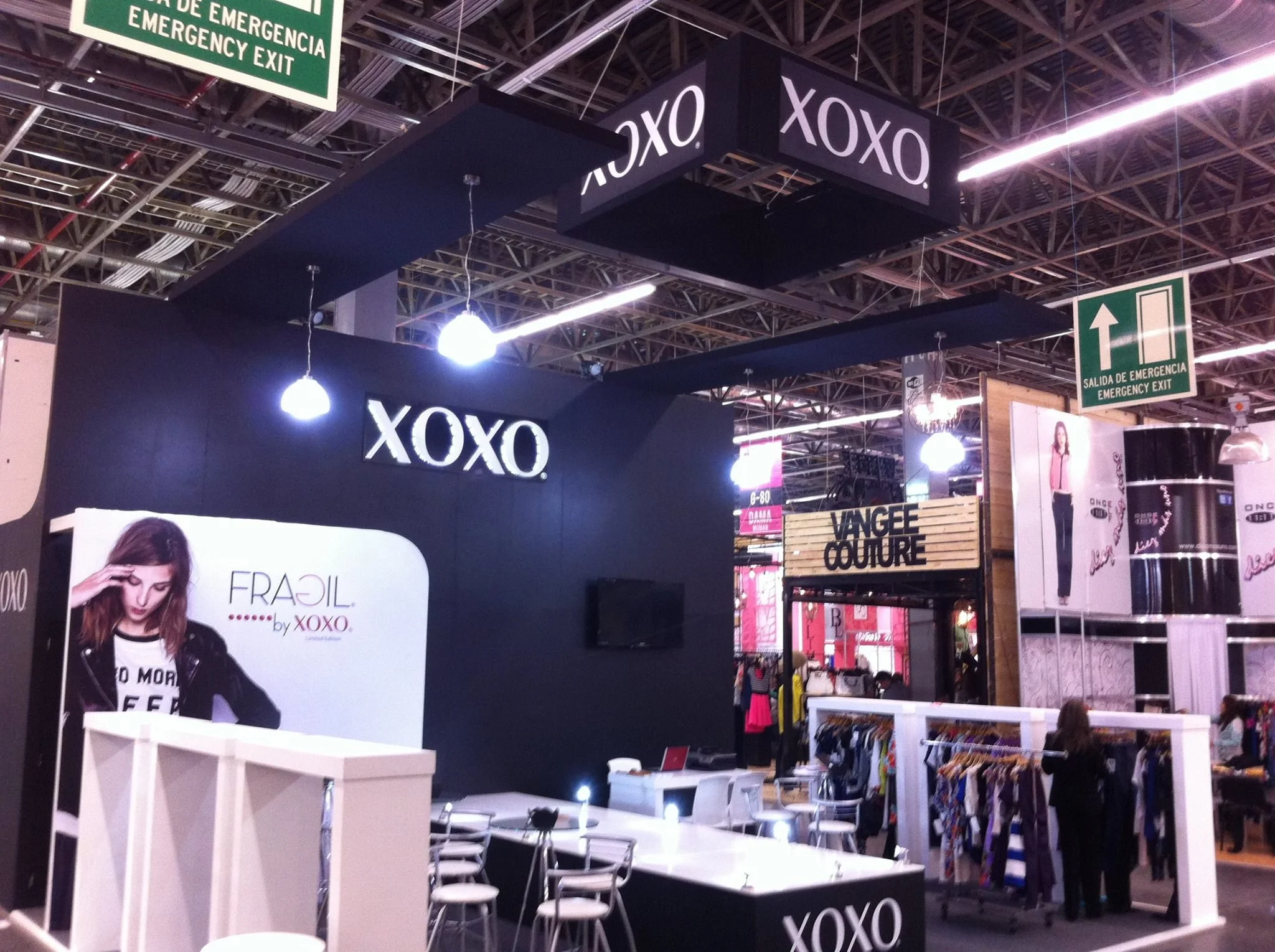 Stand de la marca XOXO en una feria comercial, con un letrero con el nombre en la parte superior y una pared negra con logo y cartel publicitario de una mujer. Se observan mesas y sillas blancas frente al puesto, además de algunas prendas en exhibici