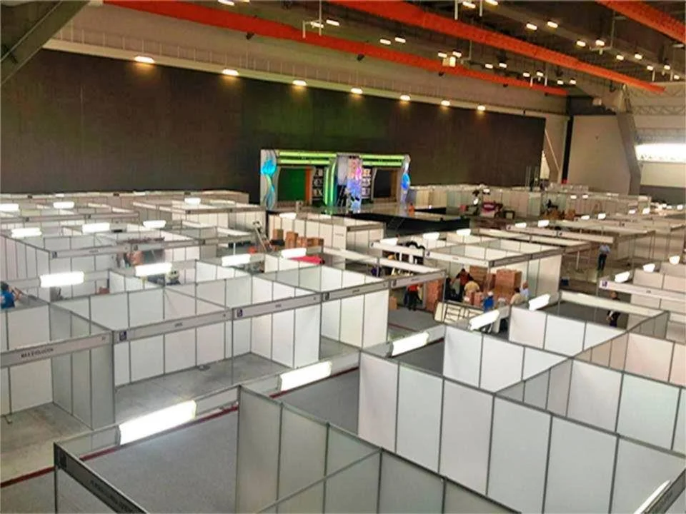 Vista de un gran salón con varias cabinas blancas, algunas con personas trabajando, y un escenario al fondo.