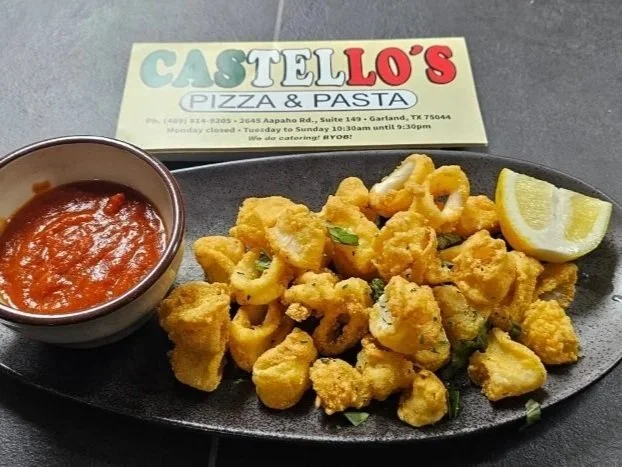 Fried calamari served with a lemon wedge and a side of marinara sauce on a black oval plate. A restaurant business card for 'Castello's Pizza & Pasta' is in the background.
