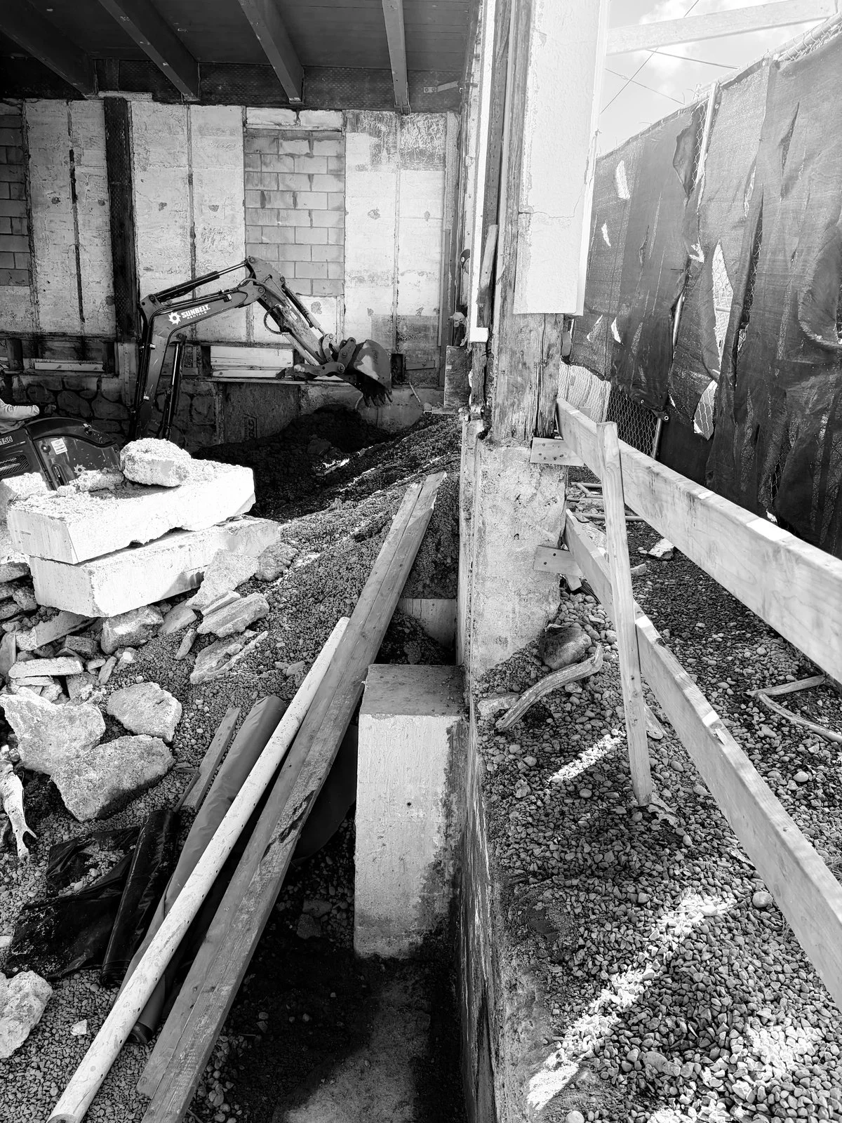 Construction site with a small excavator working inside a building, surrounded by concrete blocks, pipes, and wooden barriers.