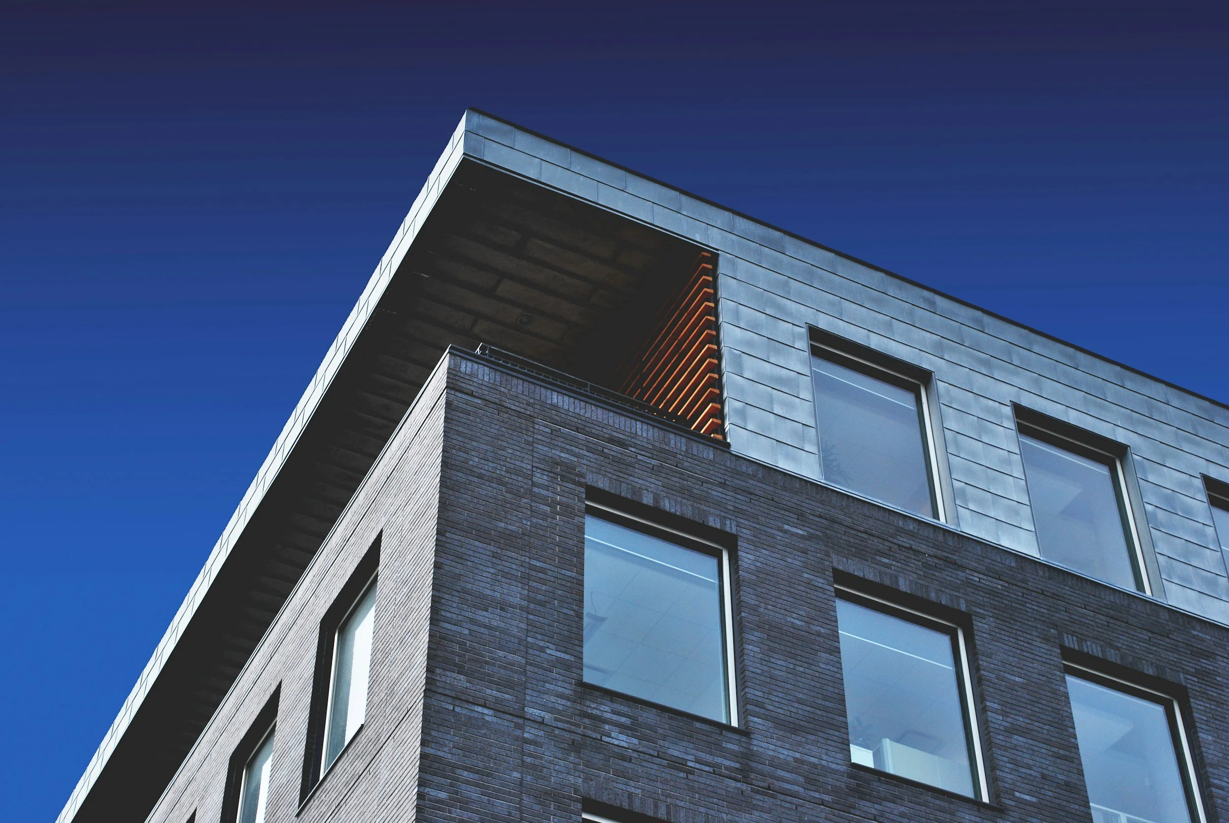 Modern multi-story building with dark brick and metal panels, large windows, under a clear blue sky.