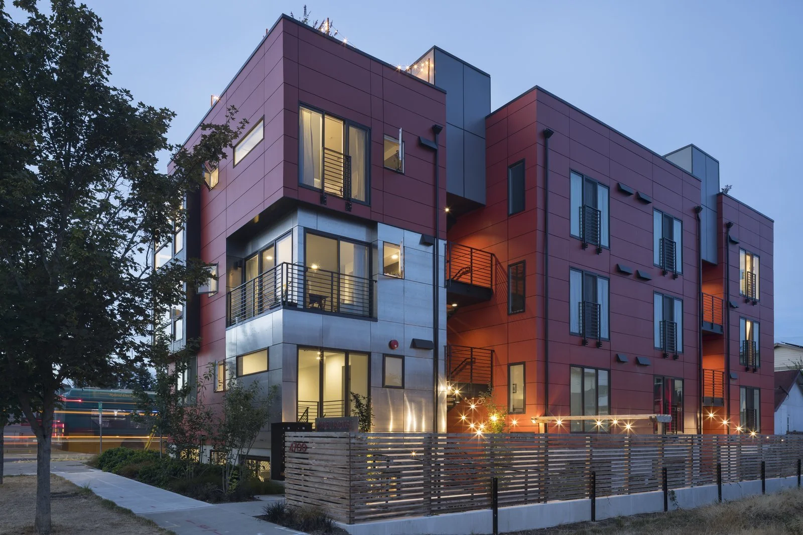 Modern multi-story apartment building at dusk with large windows, balconies, and exterior lighting, surrounded by a fence and trees.