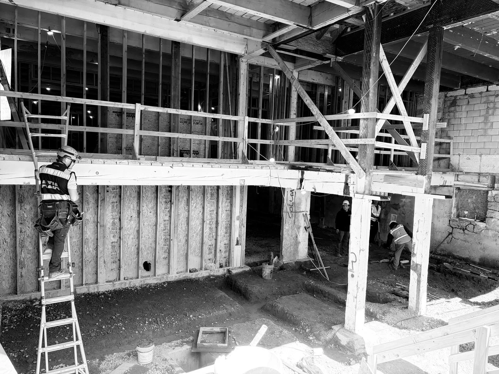 Construction workers building a multi-level structure, with one worker on a ladder in the foreground, and others working on the ground, with visible wooden framing, scaffolding, and construction tools.