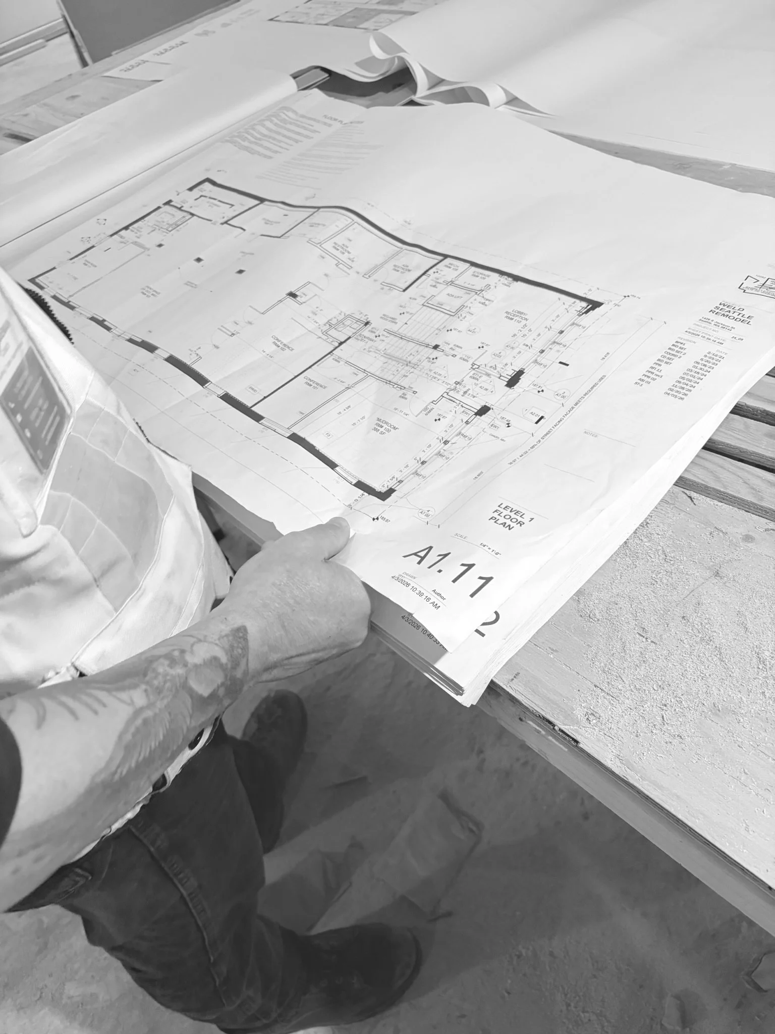 Architectural blueprint plans on a desk with a person holding a corner, showing detailed floor layouts and measurements.