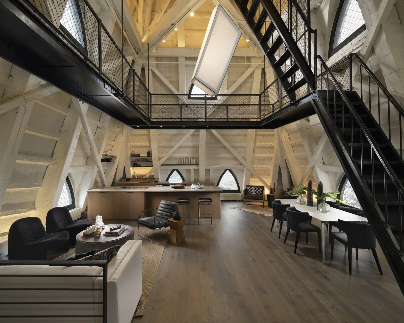 Modern loft-style interior with high vaulted ceiling, exposed white beams, and large arched windows. The space includes a kitchen area with a wooden island, a sitting area with black and white furniture, a dining table with black chairs, and a mezzanine with black metal stairs and railing.