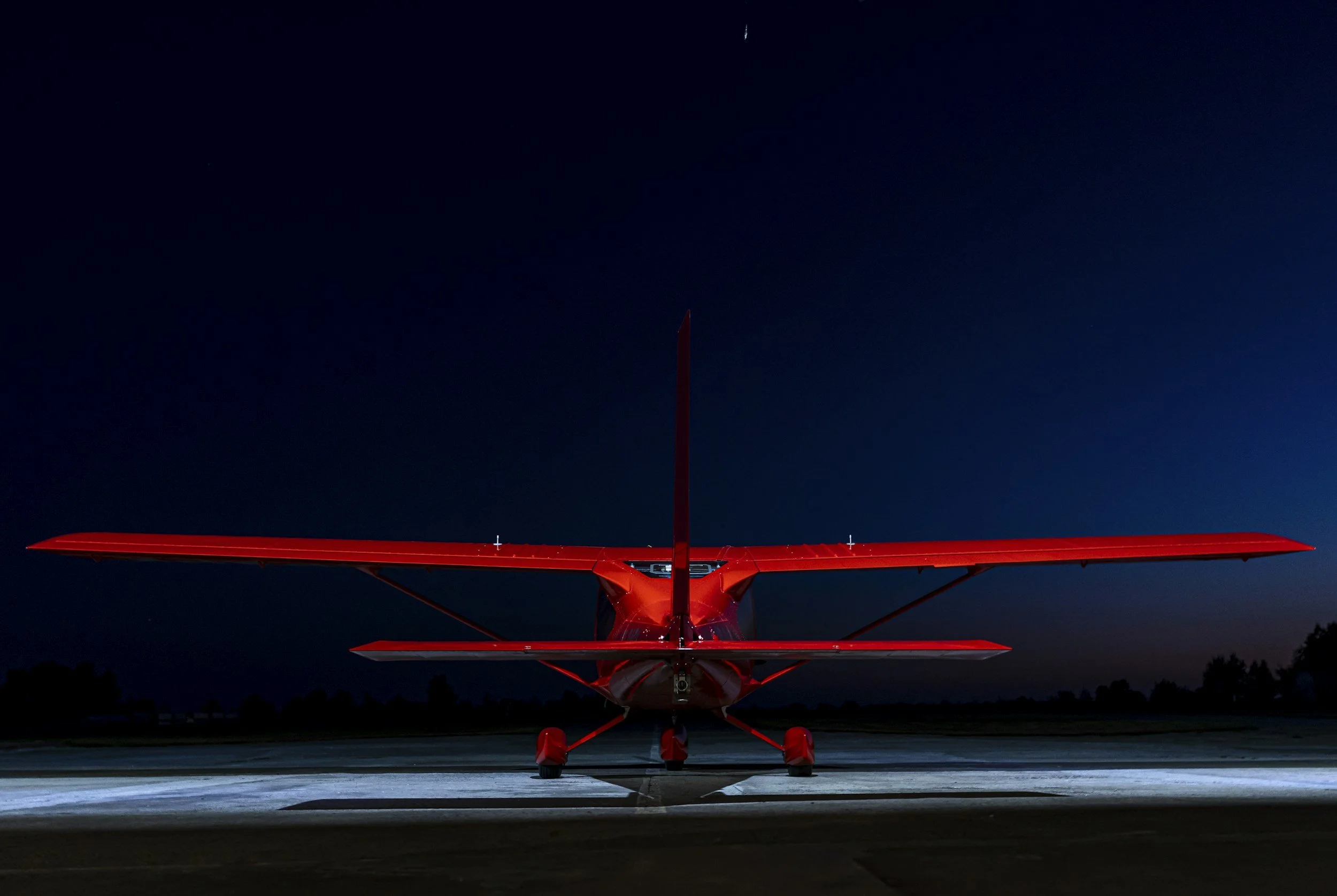Red airplane parked on the tarmac at night, illuminated by ground lights, with a dark sky and faint stars in the background.