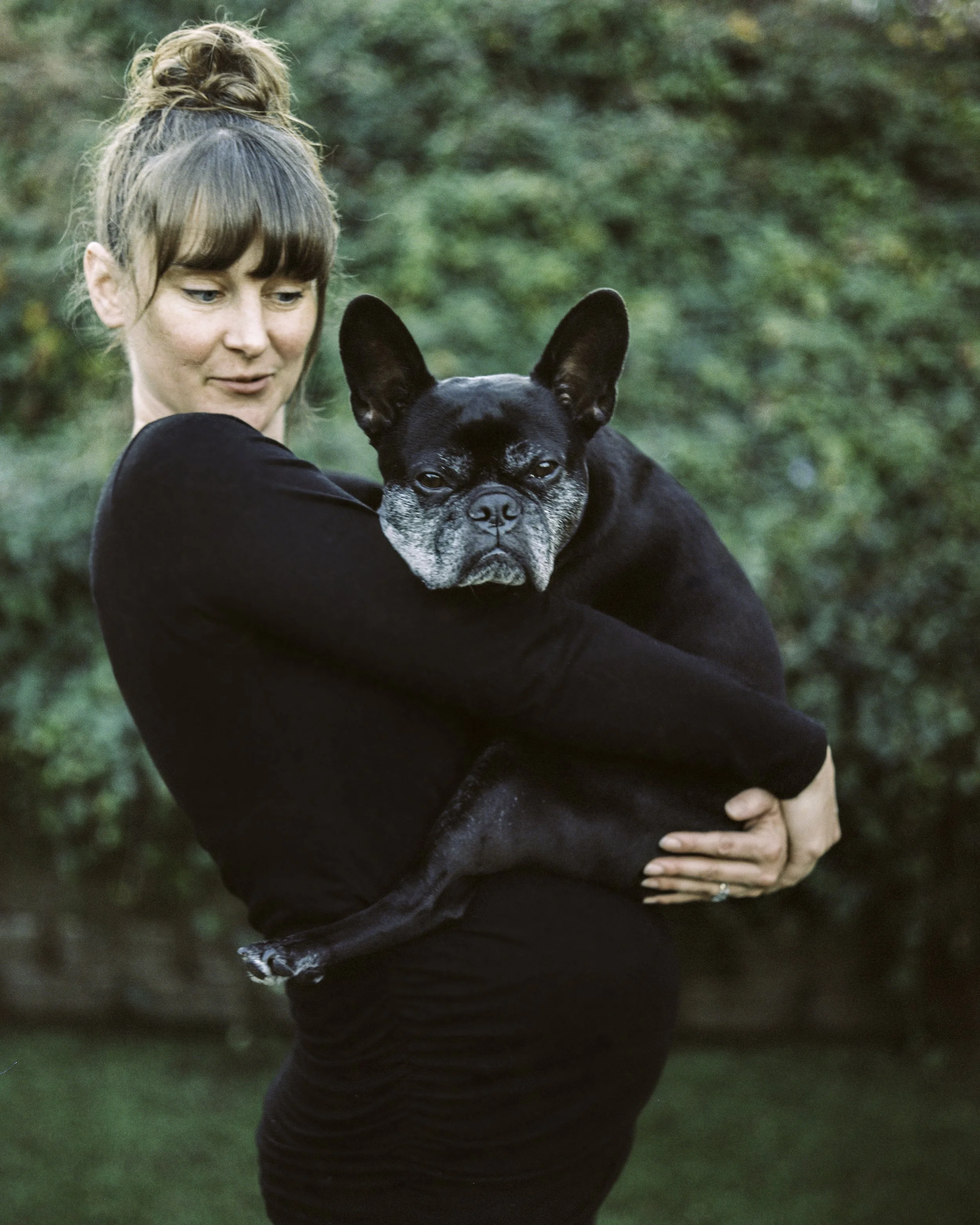 A woman with light skin and brown hair in a bun, wearing a black long-sleeve shirt, holding a black and gray French Bulldog outdoors with a blurred green background.