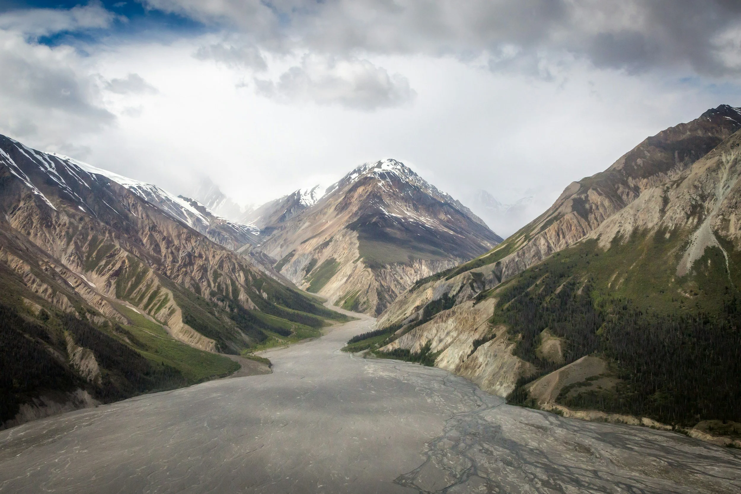 A mountain valley with steep, rugged mountains on both sides, some covered in snow, and a wide, winding dry riverbed in the foreground. Overcast sky with clouds.