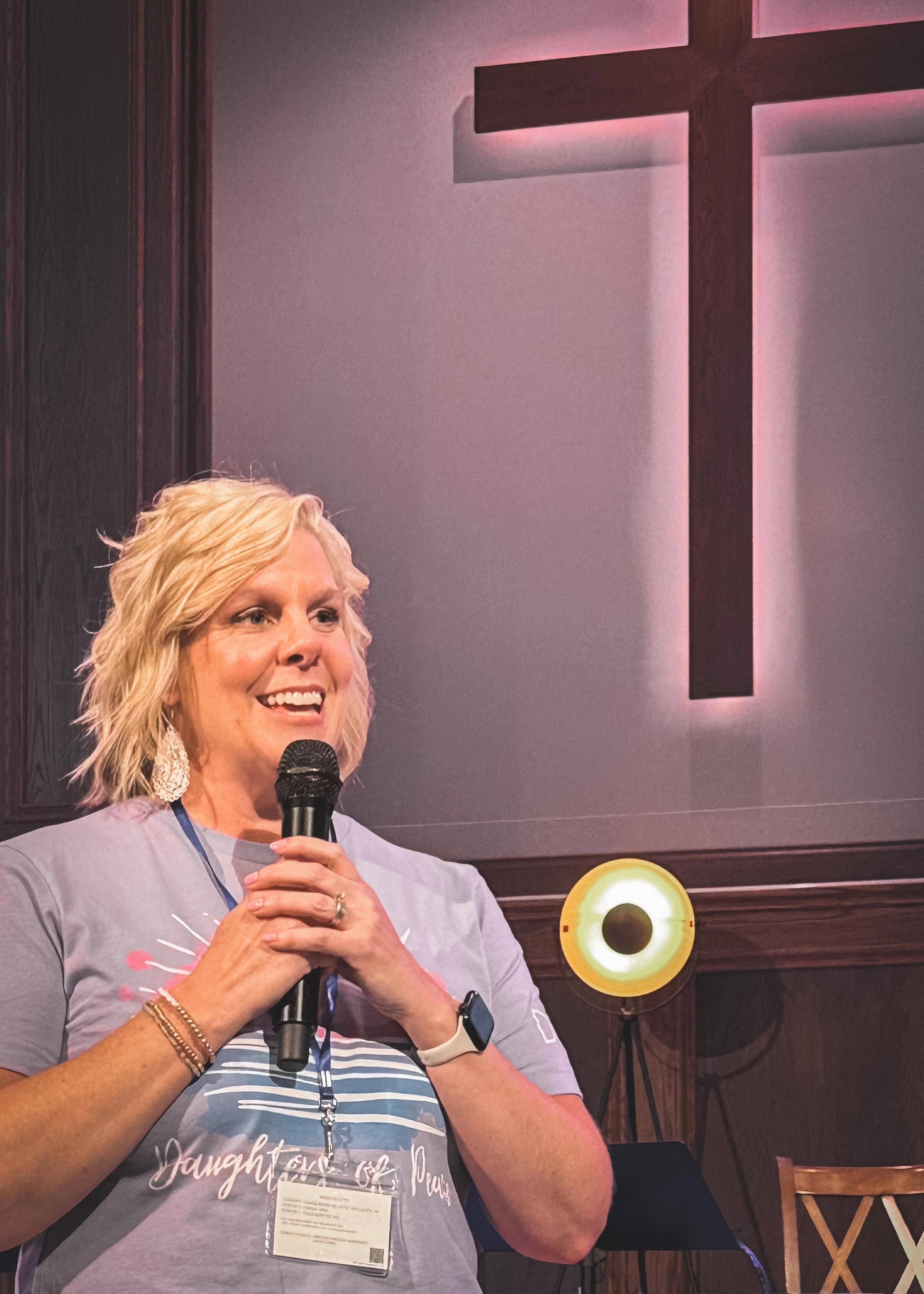 Woman with blond hair holding a microphone speaking at an indoor event, wearing a gray t-shirt, with a wooden wall and yellow circular speaker in the background.