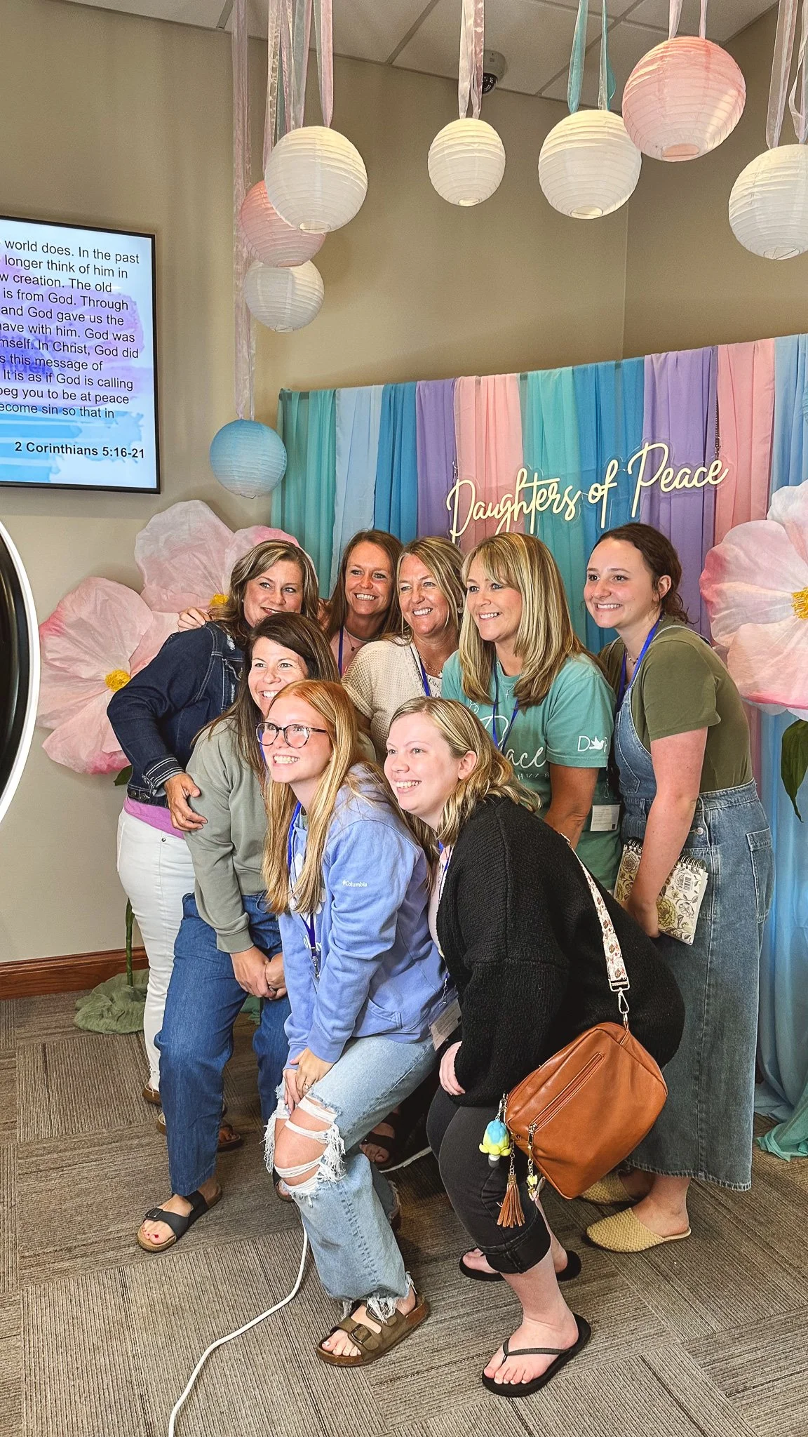 A group of women gather for a photo in front of a colorful backdrop that reads 'Daughters of Peace.' Decorations include pastel-colored paper lanterns and large pink flower cutouts on the wall. The women are smiling and dressed casually.