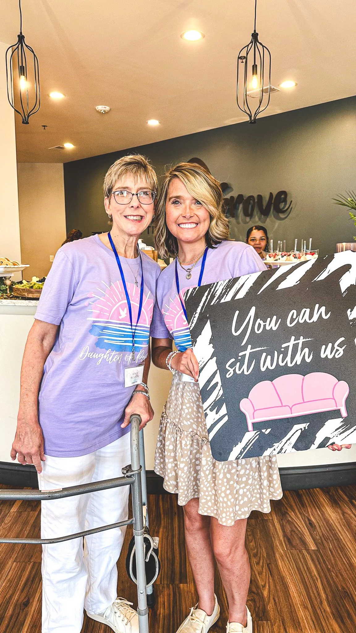 Two women standing together indoors at a social event, smiling at the camera. One is older, wearing glasses, a lavender T-shirt with graphics, and white pants, using a walker. The other woman is younger, with shoulder-length blonde hair, wearing a beige polka dot dress and holding a large sign that reads, 'You can sit with us,' with a pink sofa illustration, and wearing white sneakers.