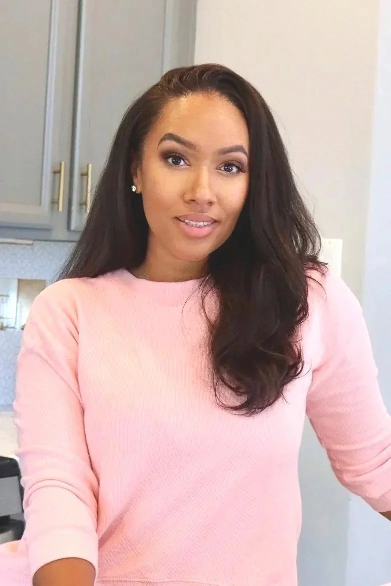 A woman with long dark hair smiling in a pink shirt in a kitchen.