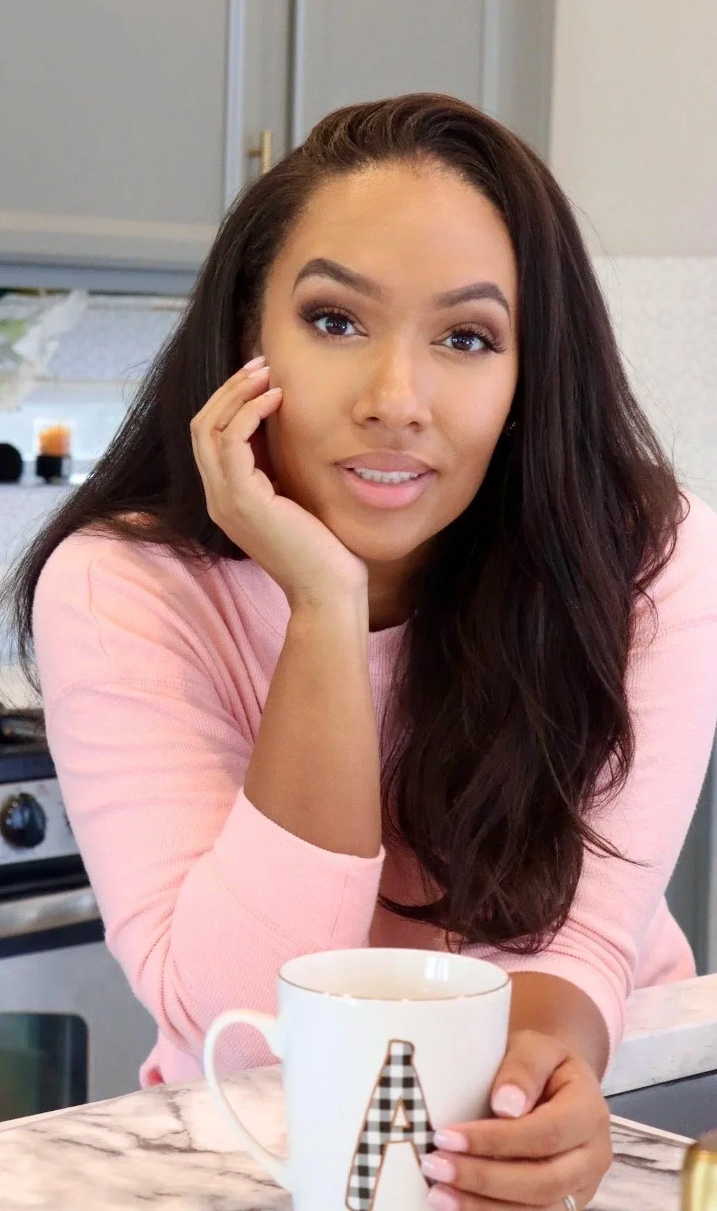 A woman with long dark hair, wearing a pink top, sitting at a kitchen counter, holding a mug with a letter A on it, smiling softly.