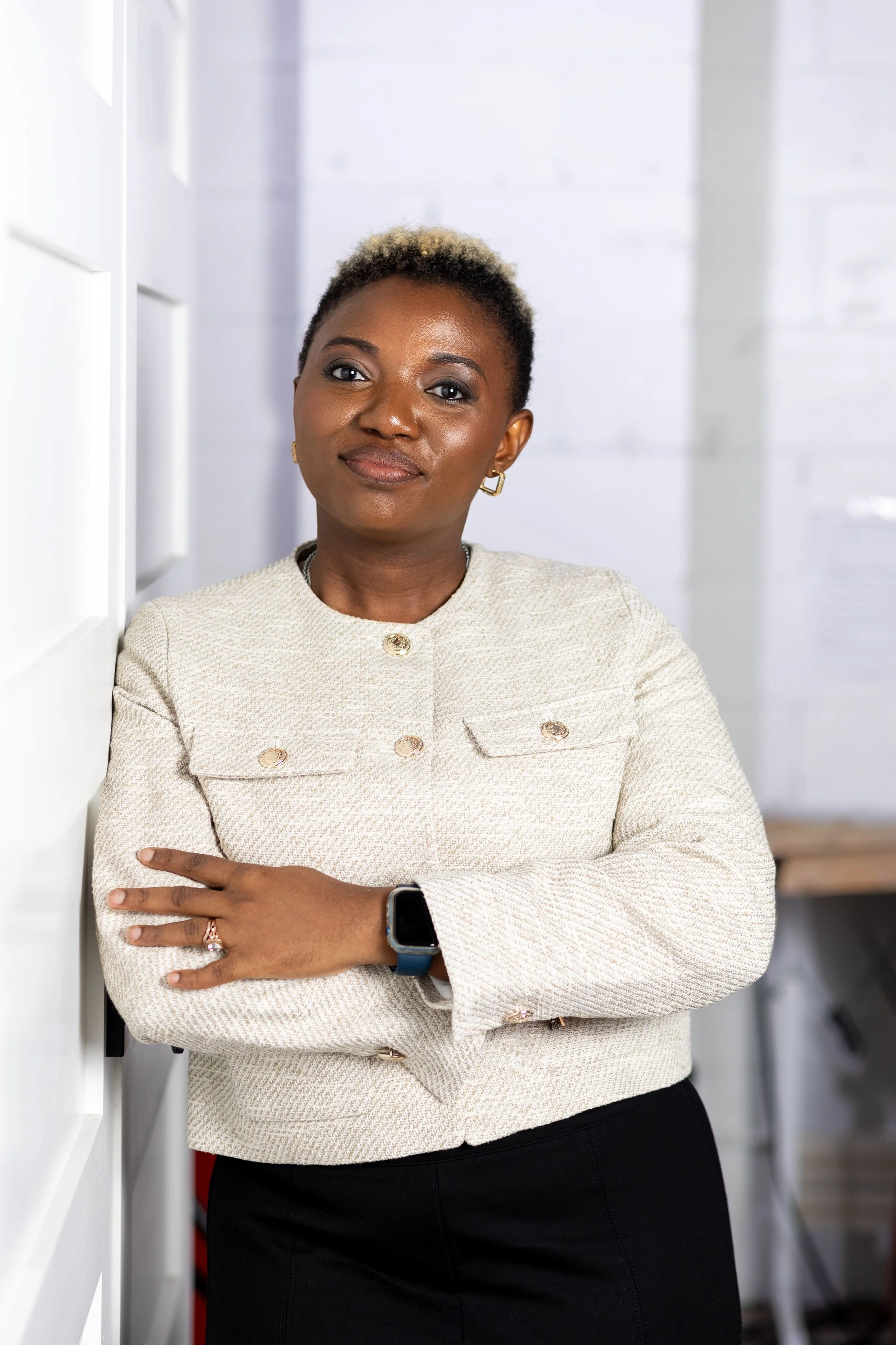A woman with short, curly hair wearing a cream-colored blazer with gold buttons, black pants, gold earrings, and a smartwatch, standing with arms crossed in front of a white wall in an office space.