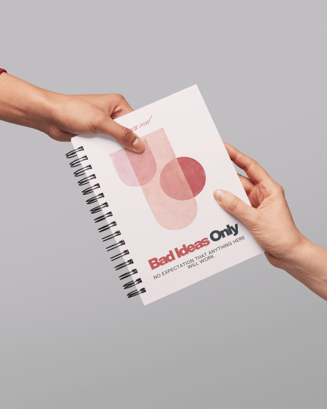 Hands exchanging a spiral notebook with the title "Bad Ideas Only" and the tagline "No expectation that anything here will work" on a gray background.
