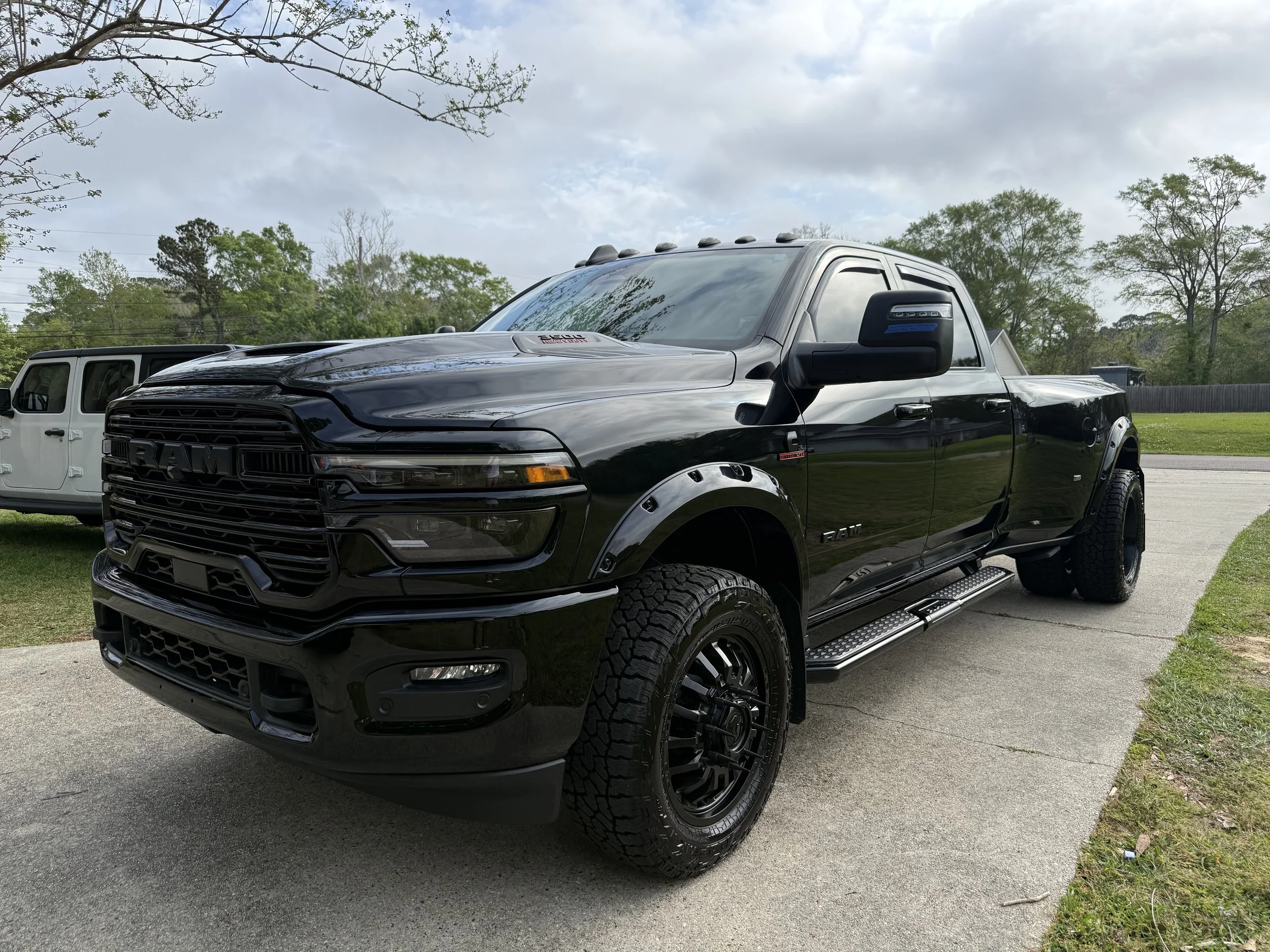 Black RAM pickup truck parked on a driveway with other vehicles, trees, and a cloudy sky in the background.