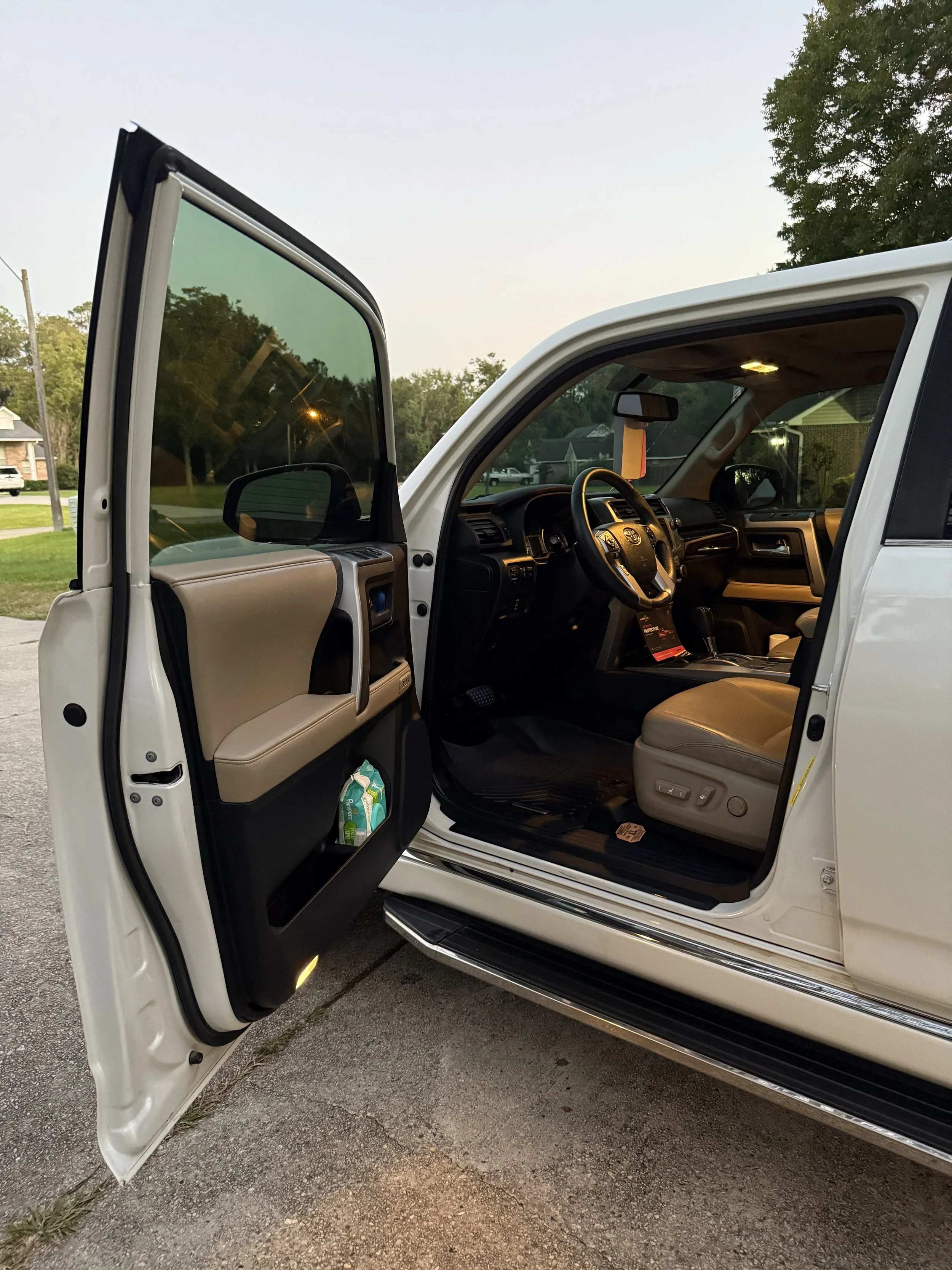 The interior view of a white pickup truck with the driver's side door open, showing the steering wheel, dashboard, and front seat with controls and a small pack of drinks in the door pocket, taken outdoors during twilight.