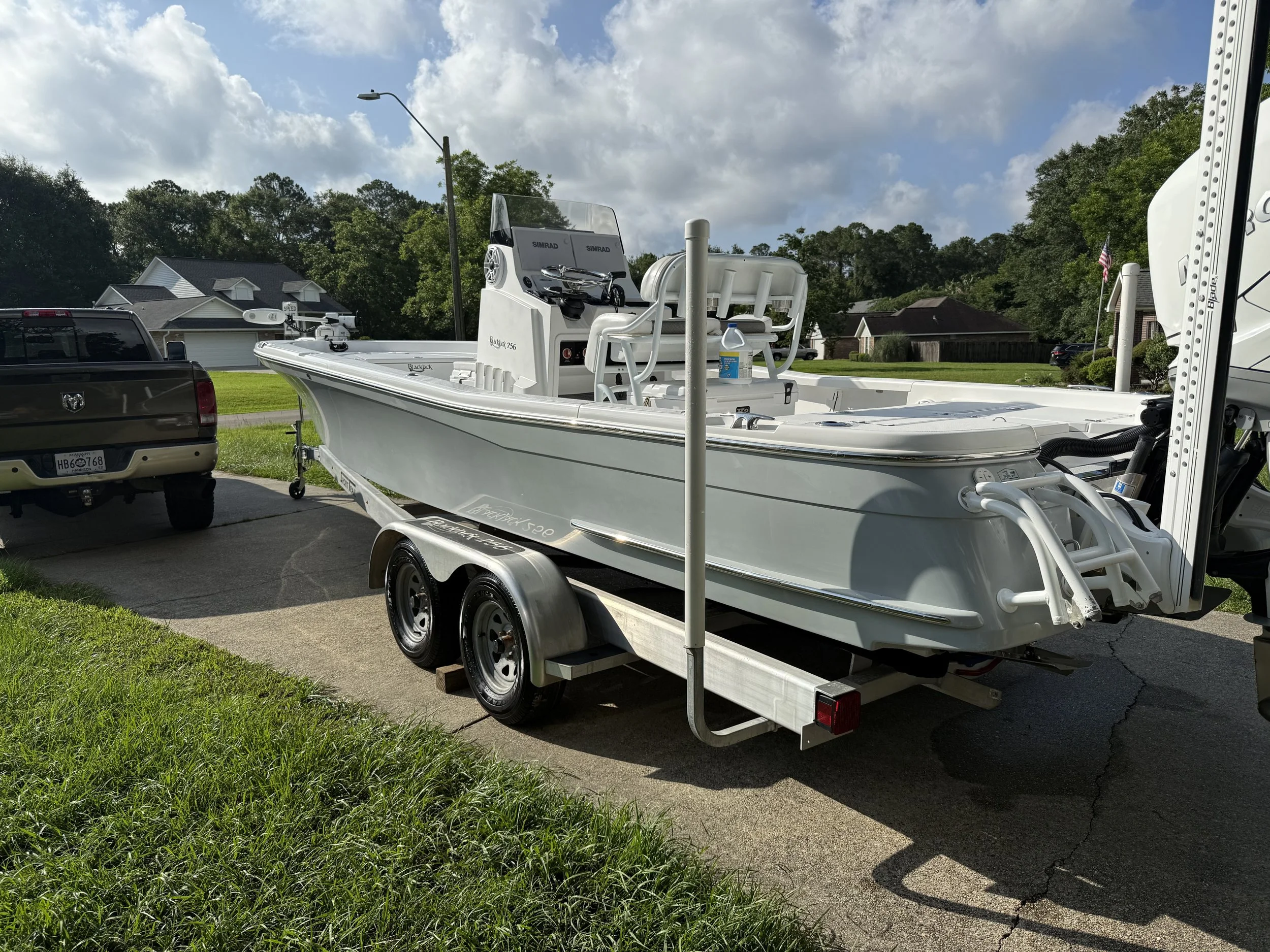 White motorboat on a trailer parked in a driveway next to a brown pickup truck, with houses and trees in the background.
