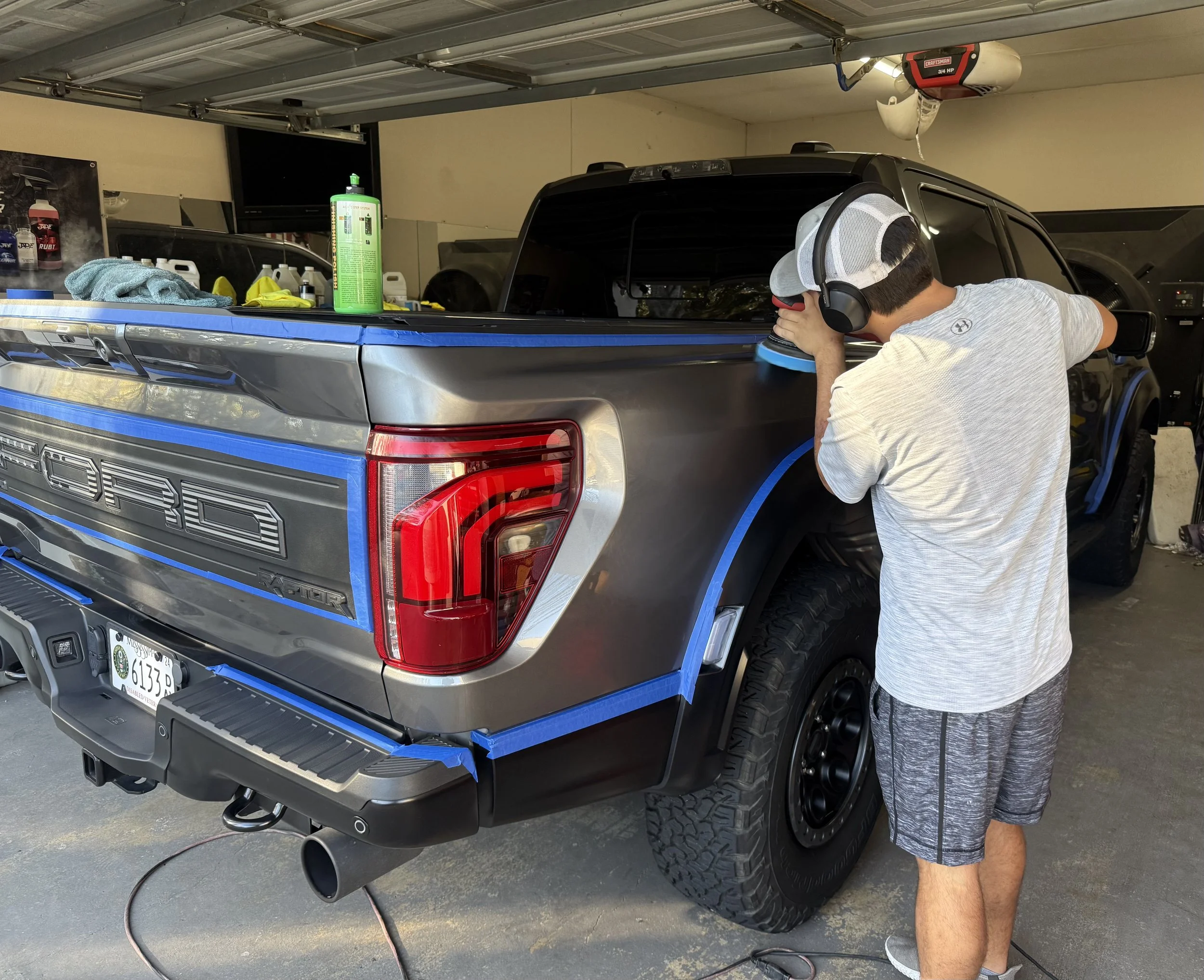 A person working on a gray Ford pickup truck in a garage, wearing headphones, a cap, and gray shorts, polishing the truck's surface with a handheld buffer.