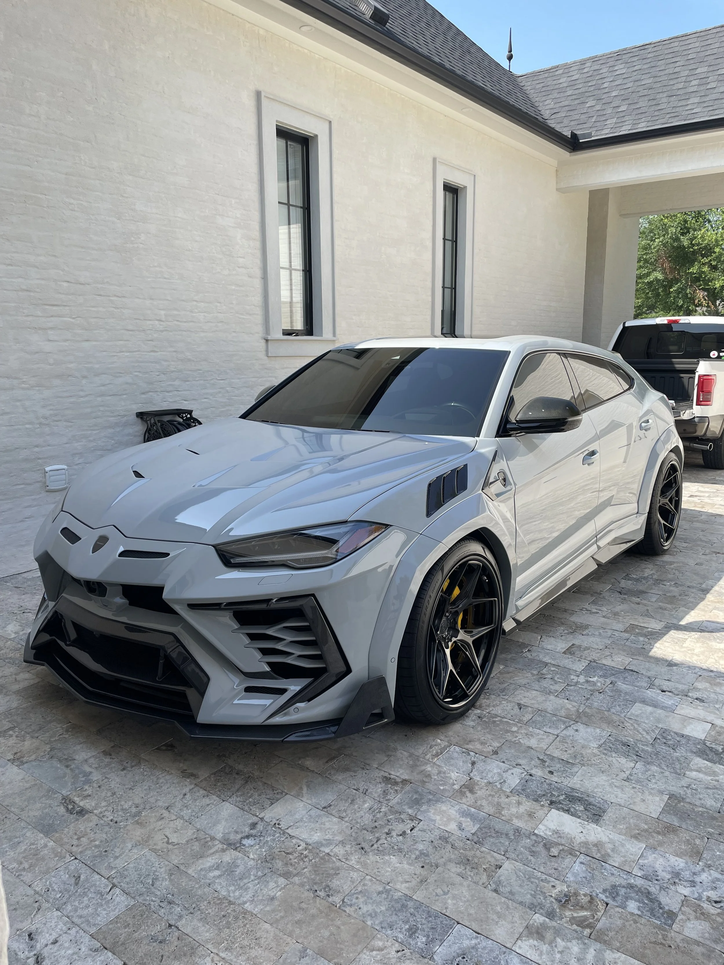 A silver Lamborghini Urus parked on a stone driveway outside a white house with tall windows and a black pickup truck in the background.