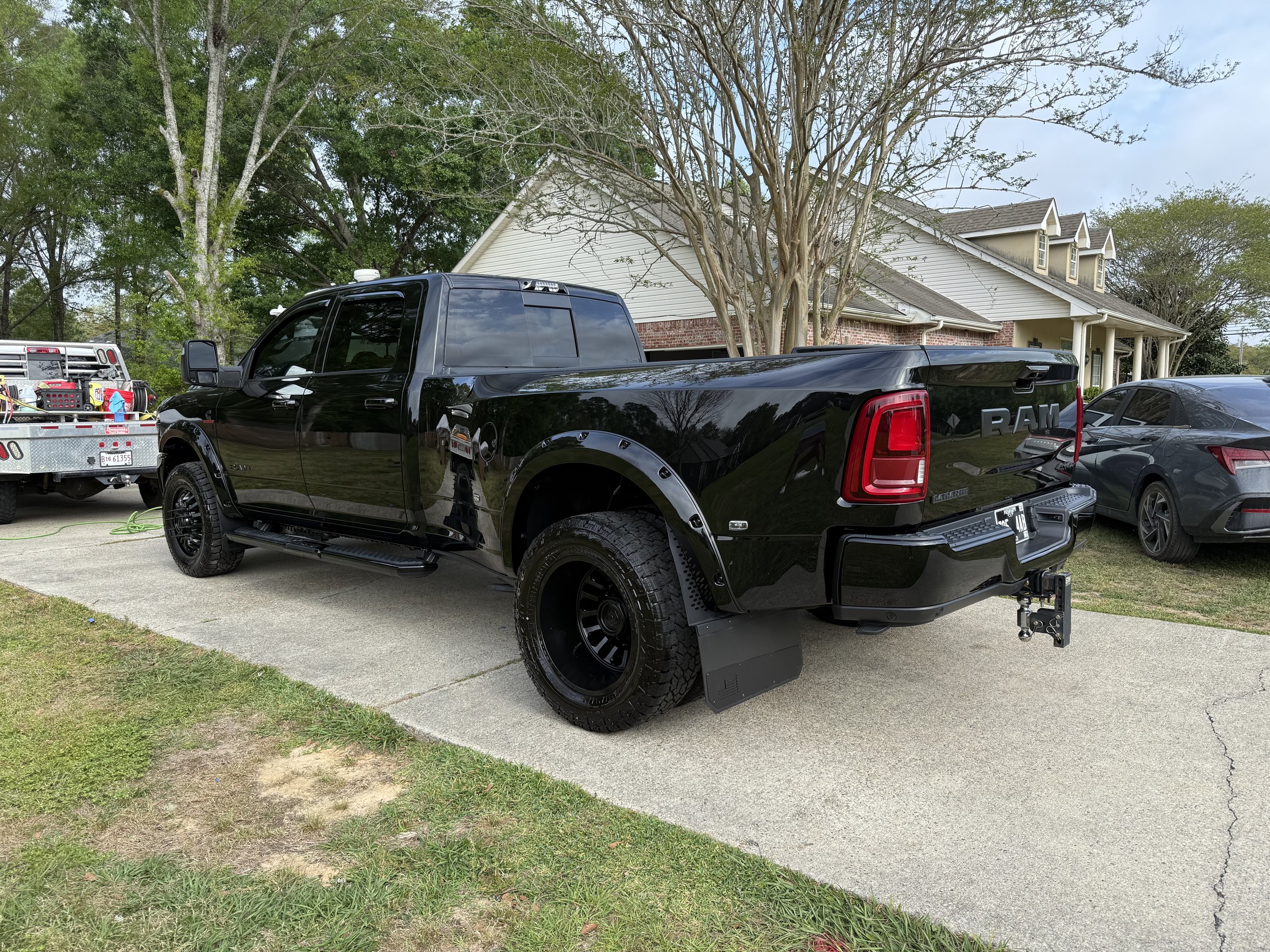 A black RAM pickup truck parked on a driveway in front of a house with a brick and white siding exterior. Trees and additional vehicles, including a gray car and a utility truck, are visible nearby.