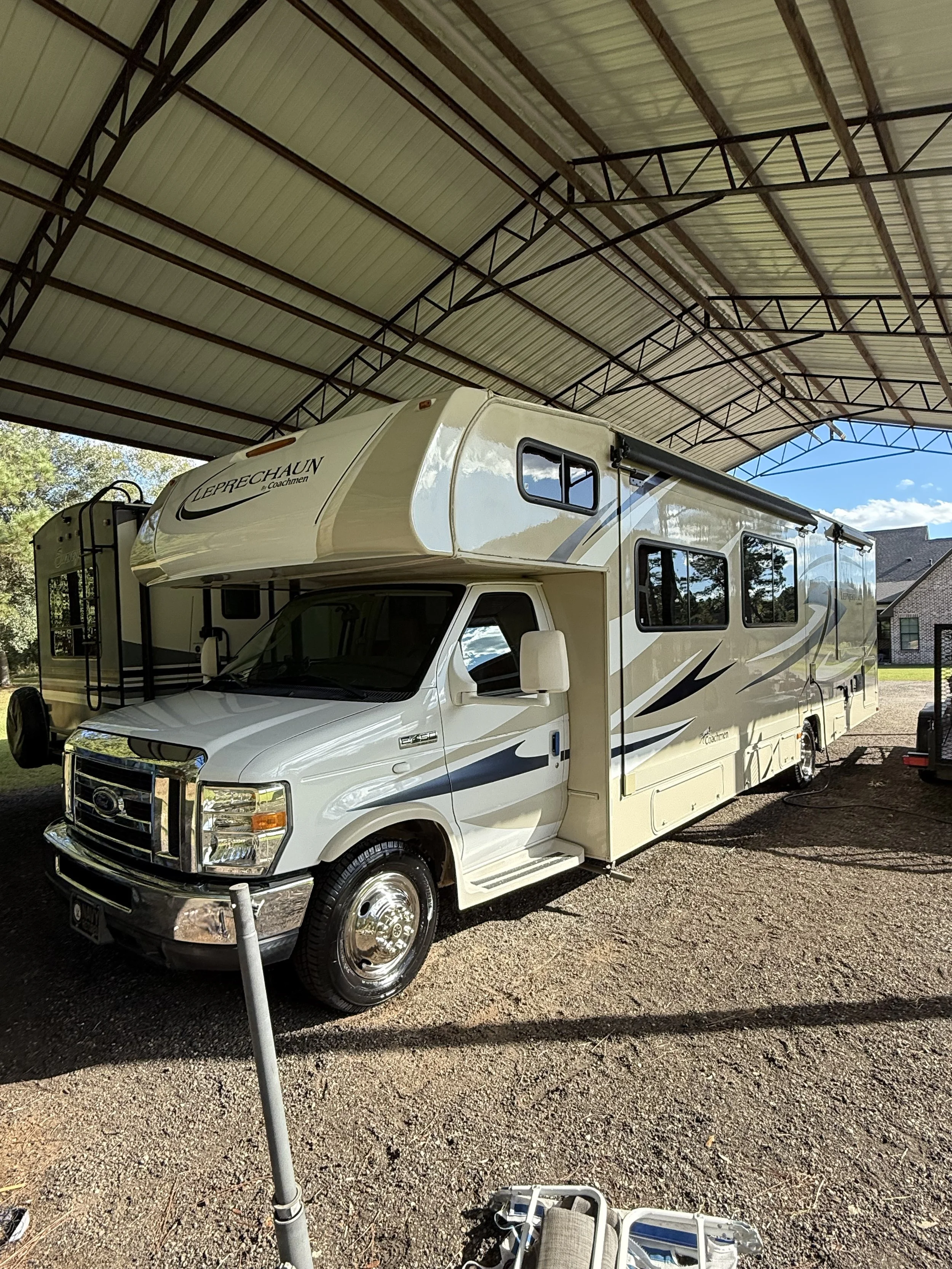 A white motorhome with black and gold accents parked under a metal canopy in a backyard. The motorhome has an over-cab bed and multiple windows, with another RV partially visible in the background.