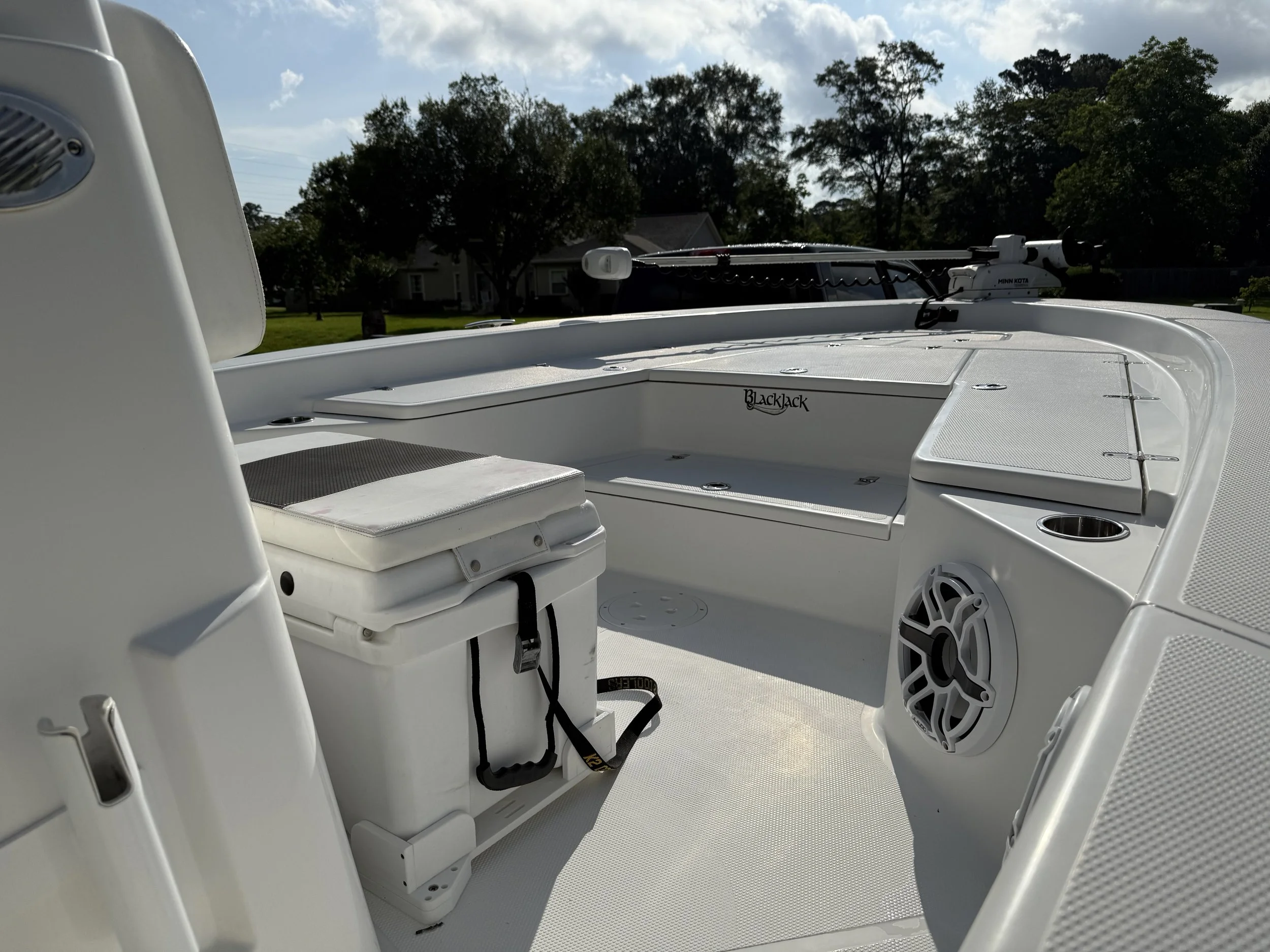The interior of a white boat with a storage box, a speaker, a cup holder, and seating area, with trees and houses in the background.