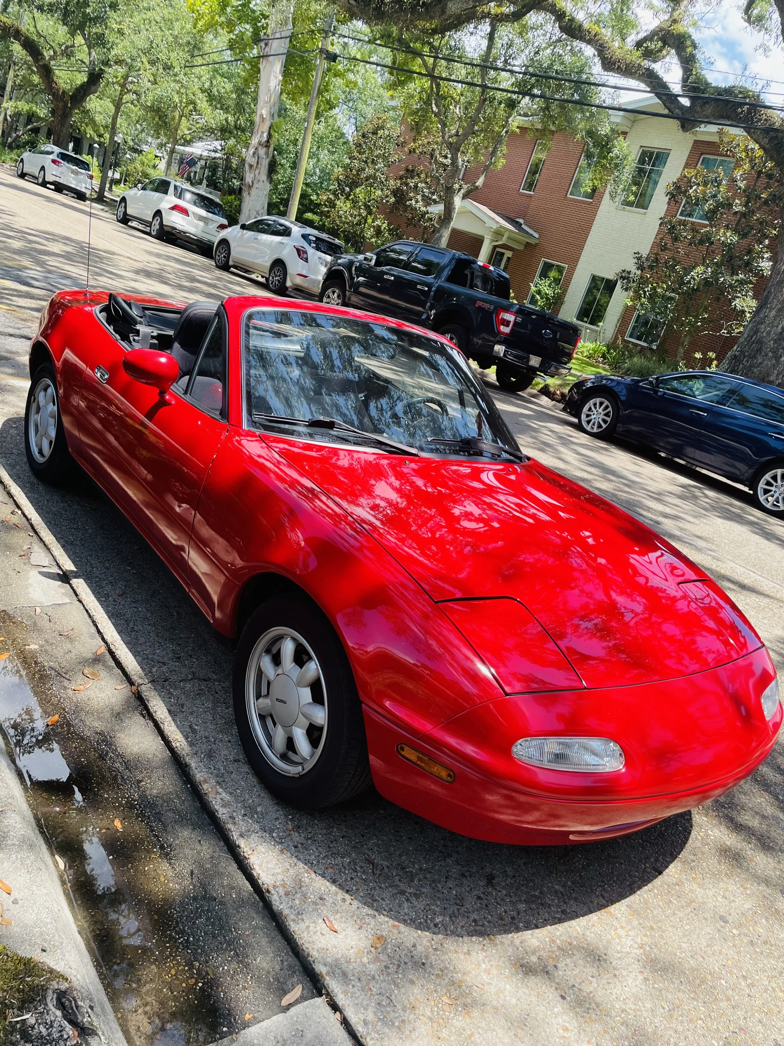 A red convertible sports car parked on the side of a residential street with other cars, trees, and houses in the background.