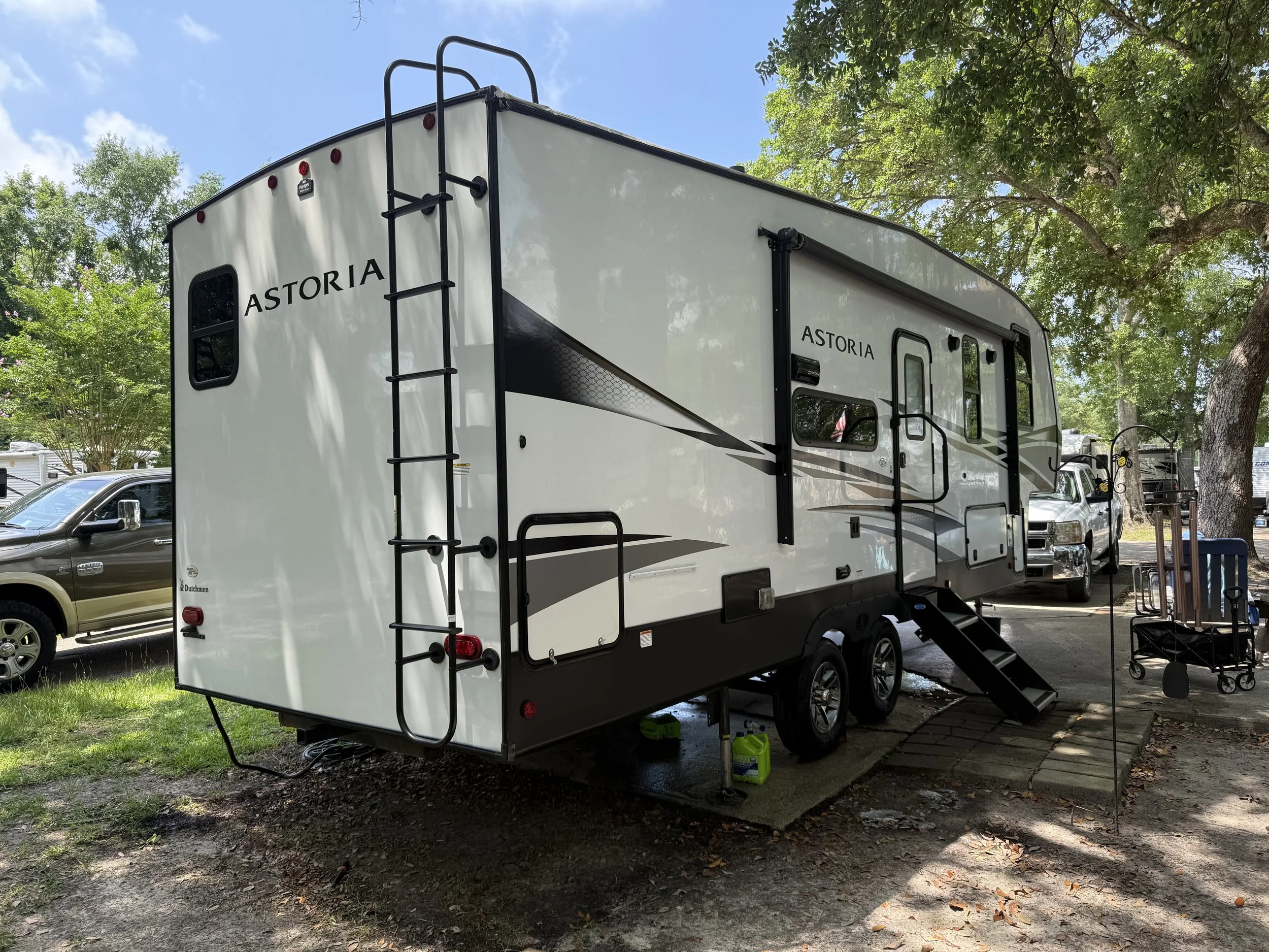 A white and black Astoria travel trailer parked in a campground with trees, cars, and camping equipment nearby.