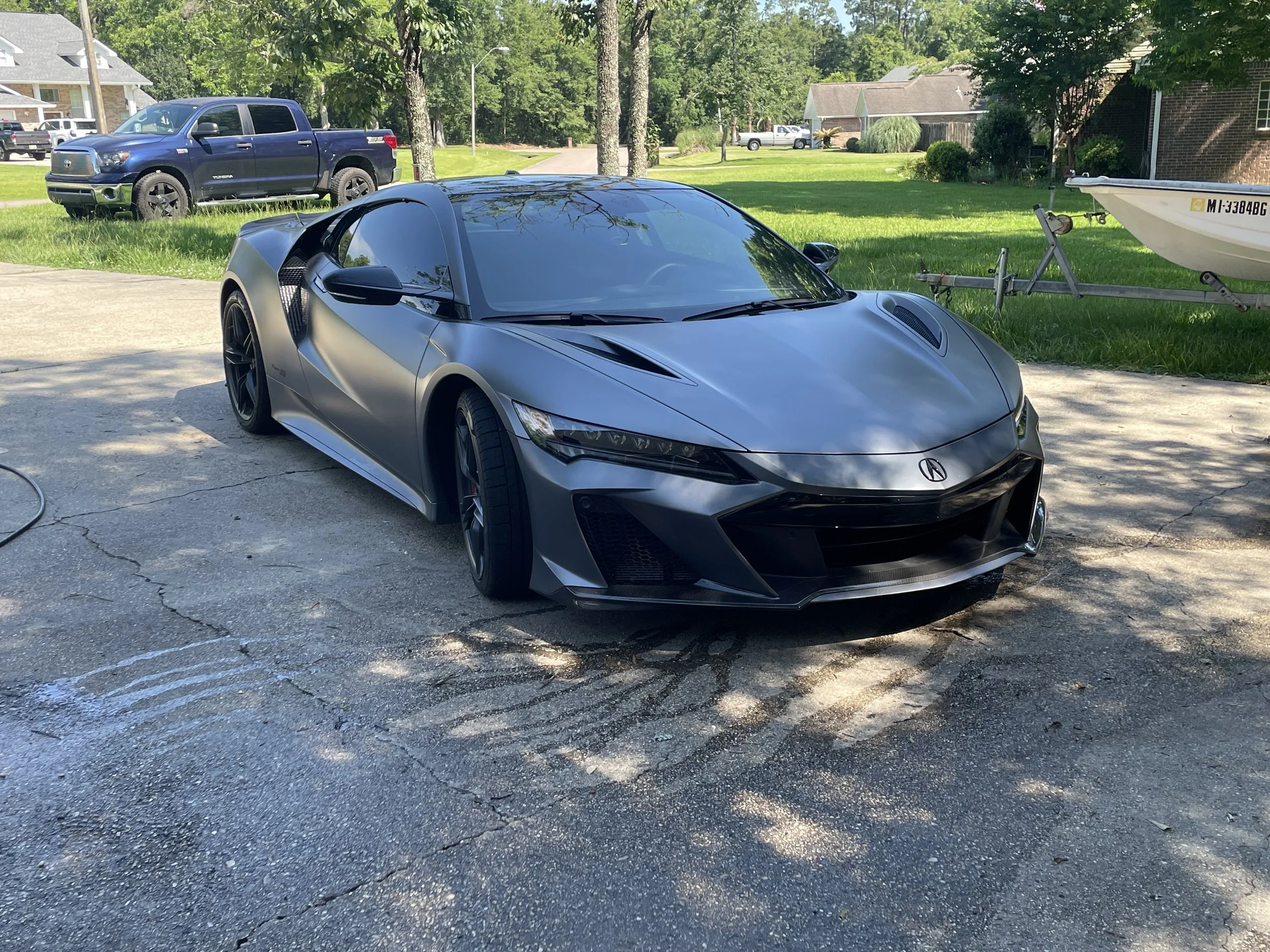 Gray sports car parked on a driveway with trees, a truck, a boat trailer, and houses in the background.