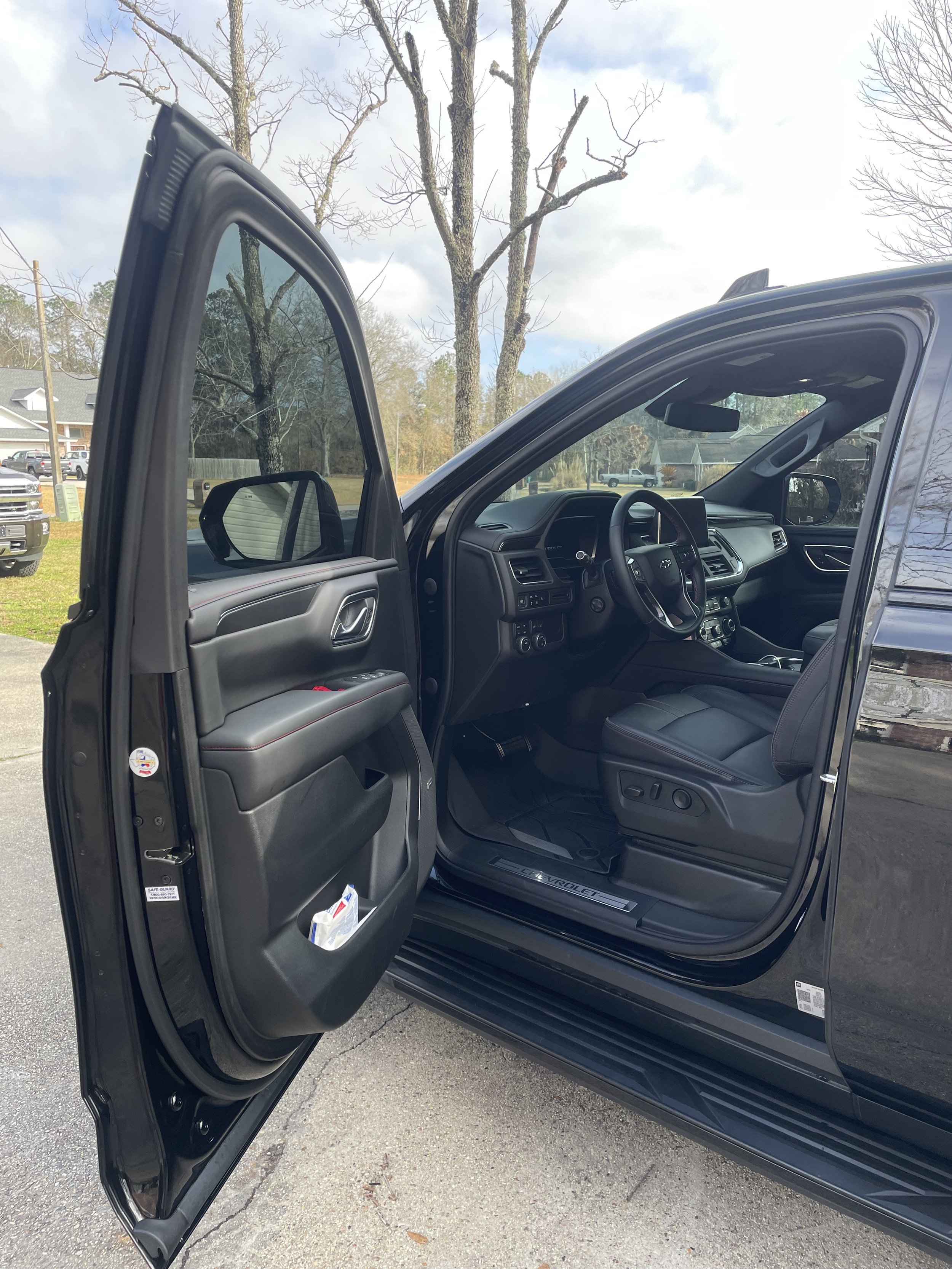 Black pickup truck with open front driver's side door, showing black leather seats and dashboard inside; outdoor parking lot with trees, cars, and cloudy sky in the background.