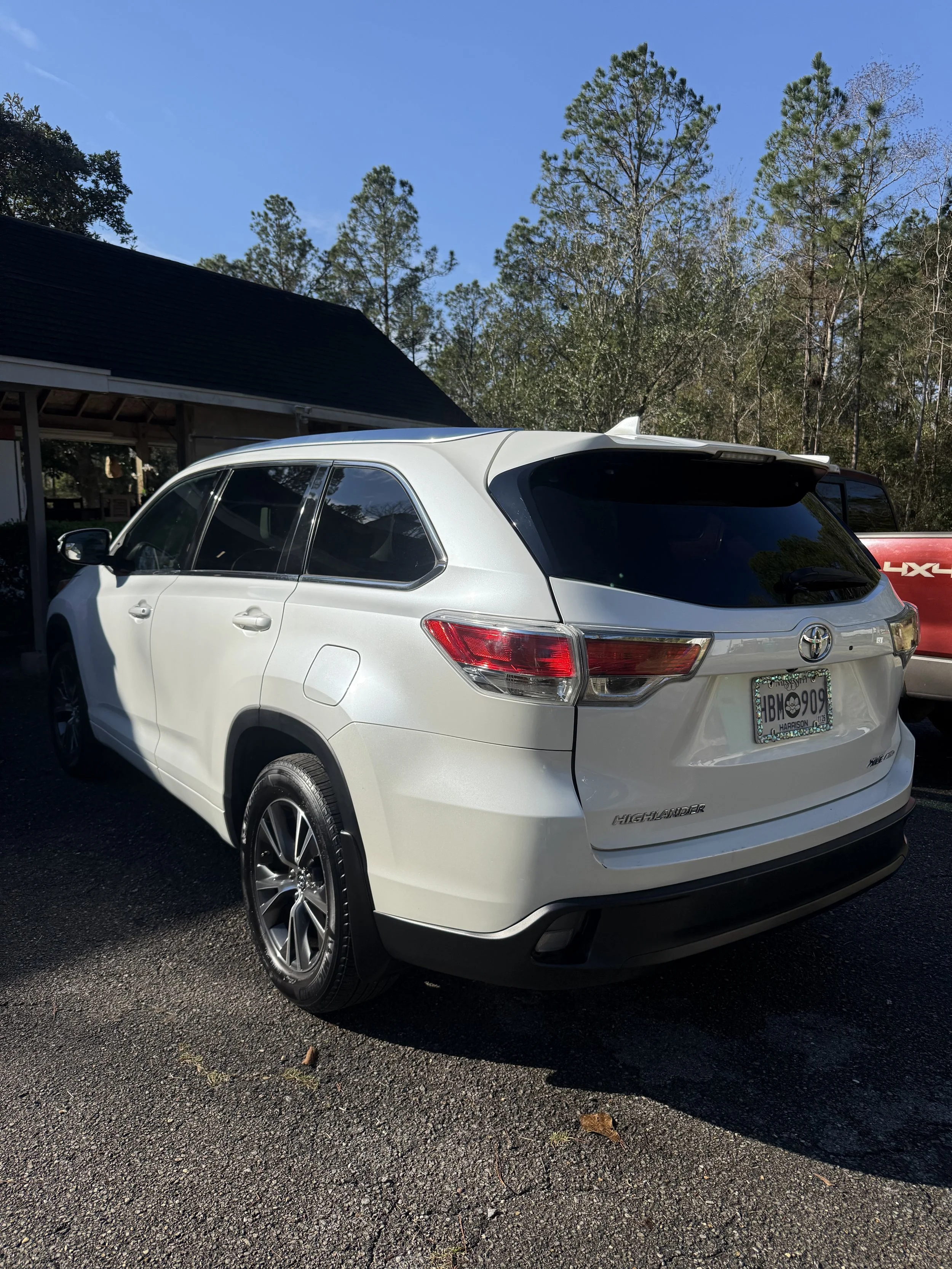 White Toyota Highlander parked outdoors on a paved surface with trees and a blue sky in the background.