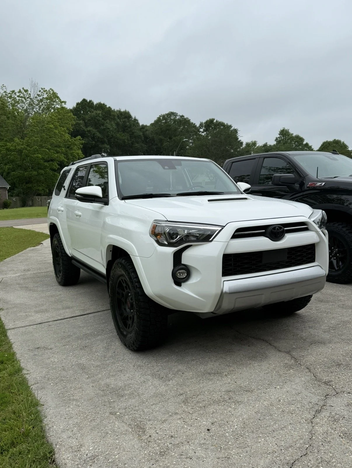 A white Toyota SUV parked on a driveway with a black pickup truck behind it and green trees in the background.
