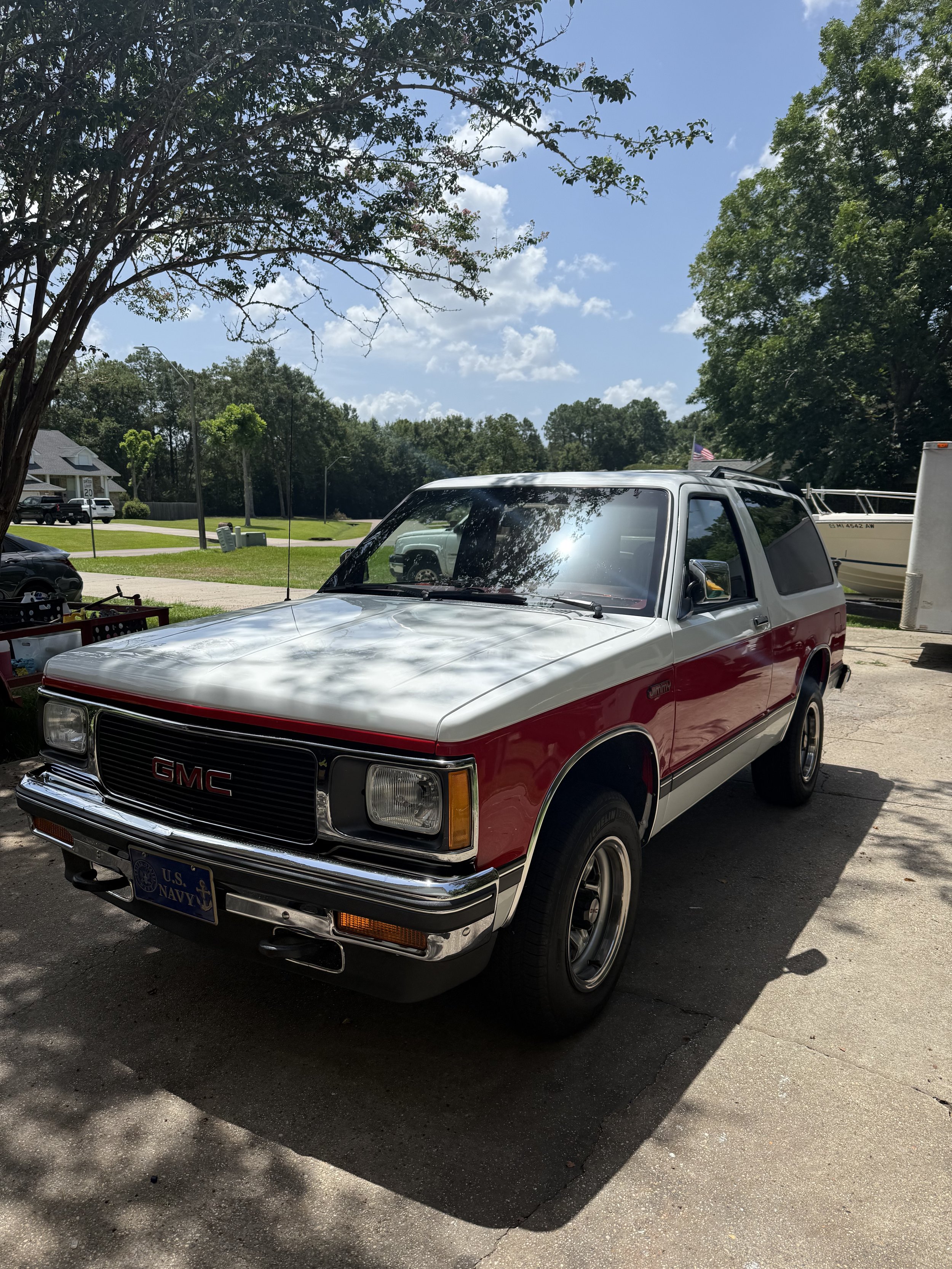 A white and red GMC SUV parked in a driveway, with a U.S. Navy license plate, under a tree with shadows cast on it, and a boat in the background.