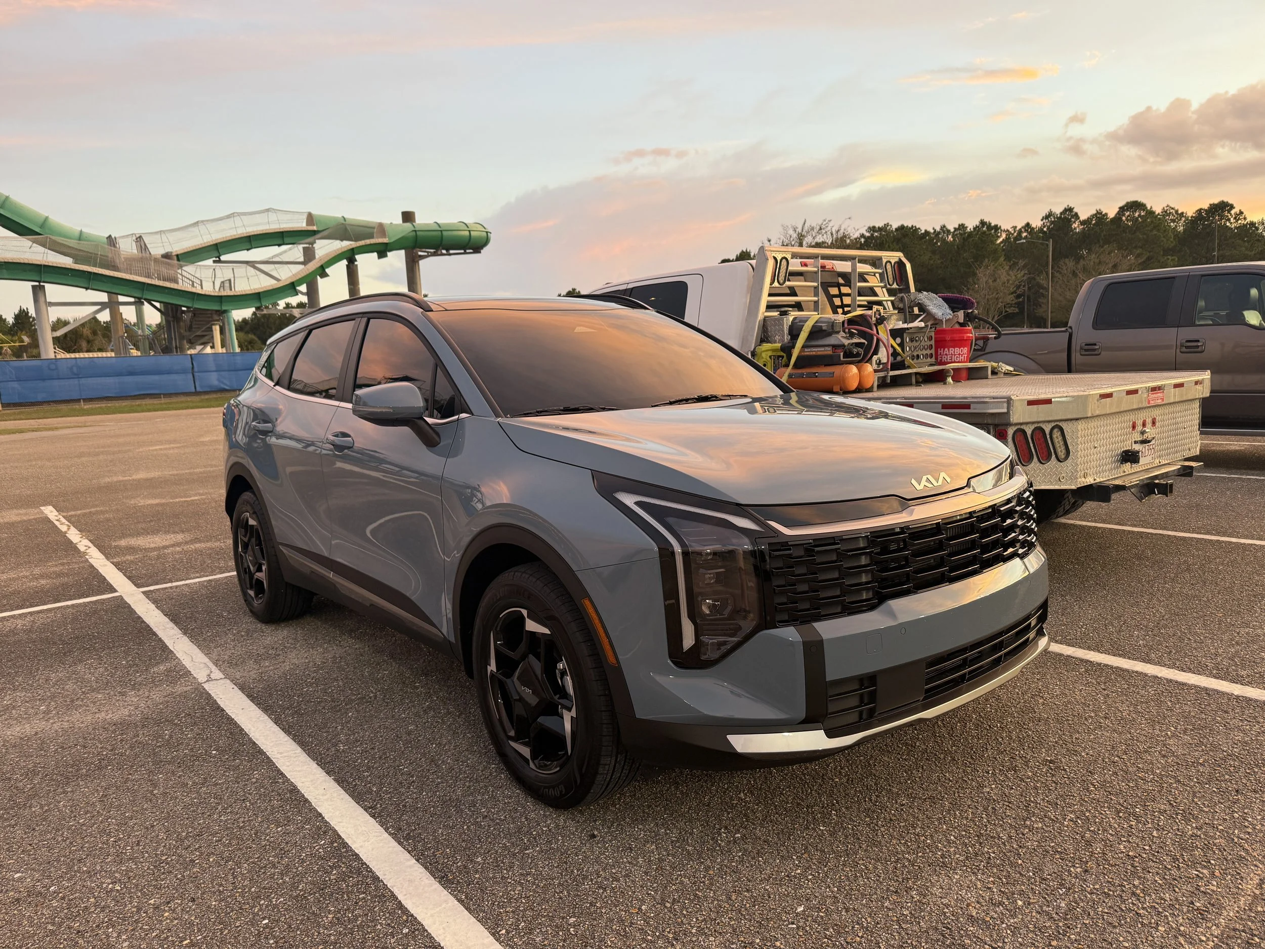 A parking lot near a water park with a gray SUV in the foreground and a trailer loaded with equipment behind it. In the background, a water slide and a blue water park barrier are visible. The sky is partly cloudy with the sun setting.