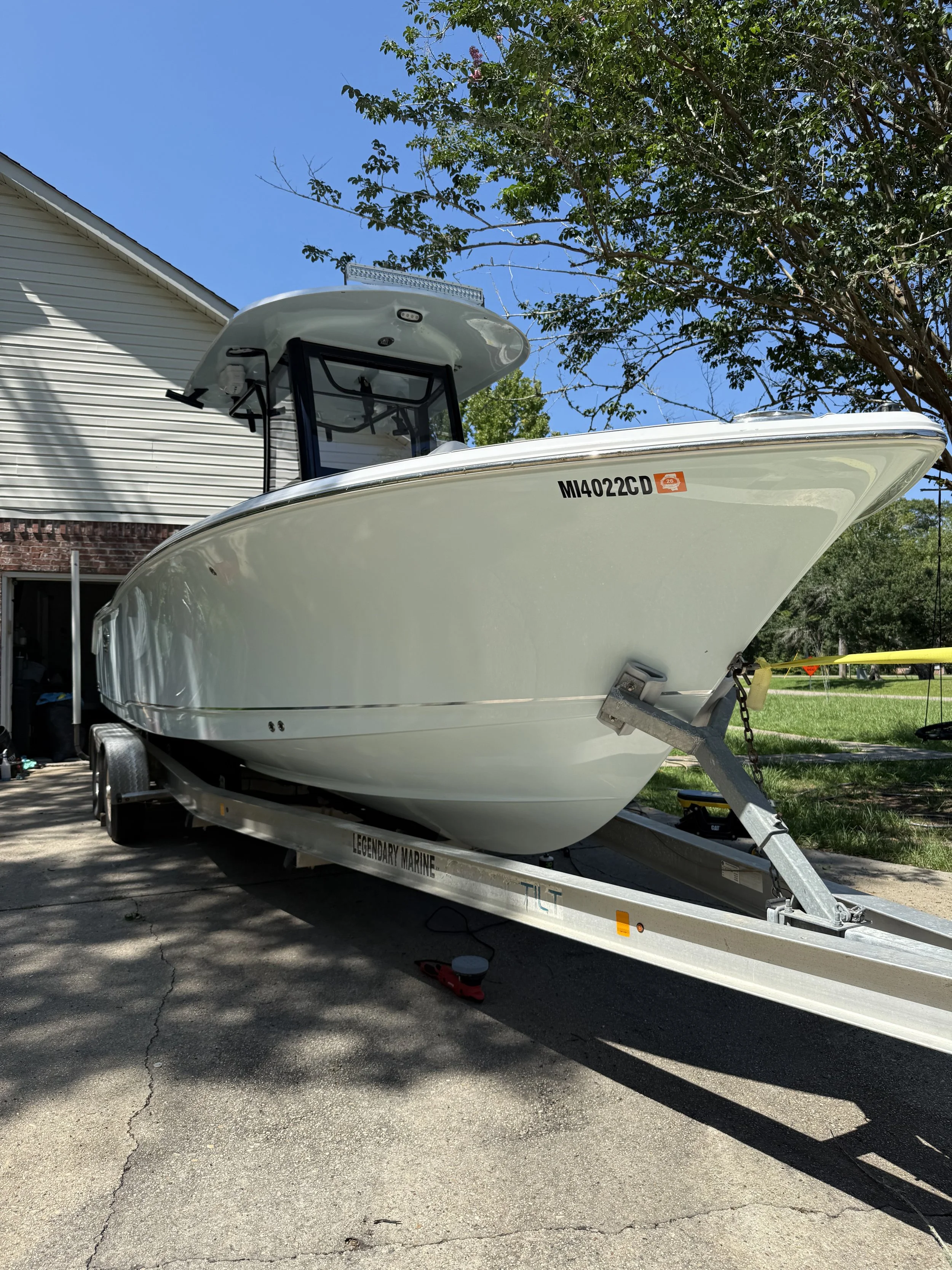 White boat on a trailer parked in a driveway with a house and trees in the background.