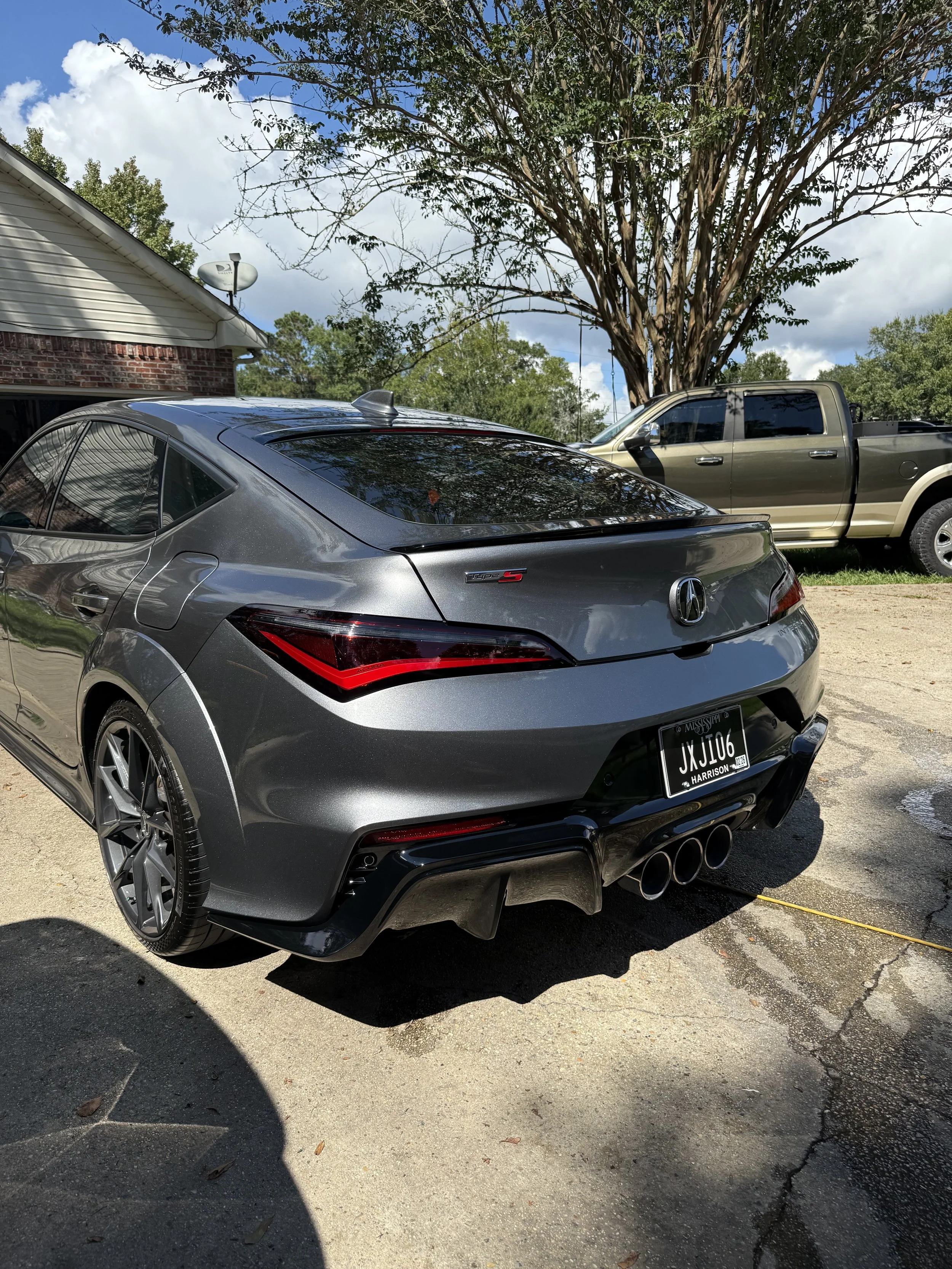 A gray Acura sports car with a black rear spoiler and quad exhaust pipes parked on a driveway, with a brown pickup truck parked on grass behind it, a large tree, a partly cloudy blue sky, and a house with a brick and white siding wall in the backgrou