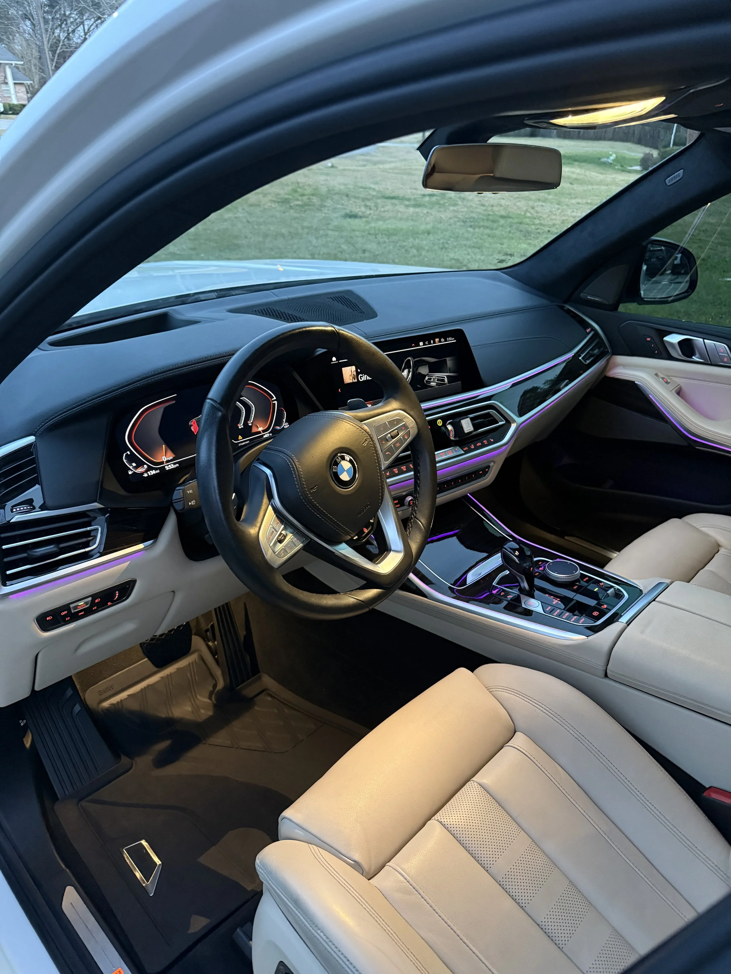 Interior of a BMW vehicle showing dashboard, steering wheel, gear shift, center console, and beige leather seats with ambient lighting.